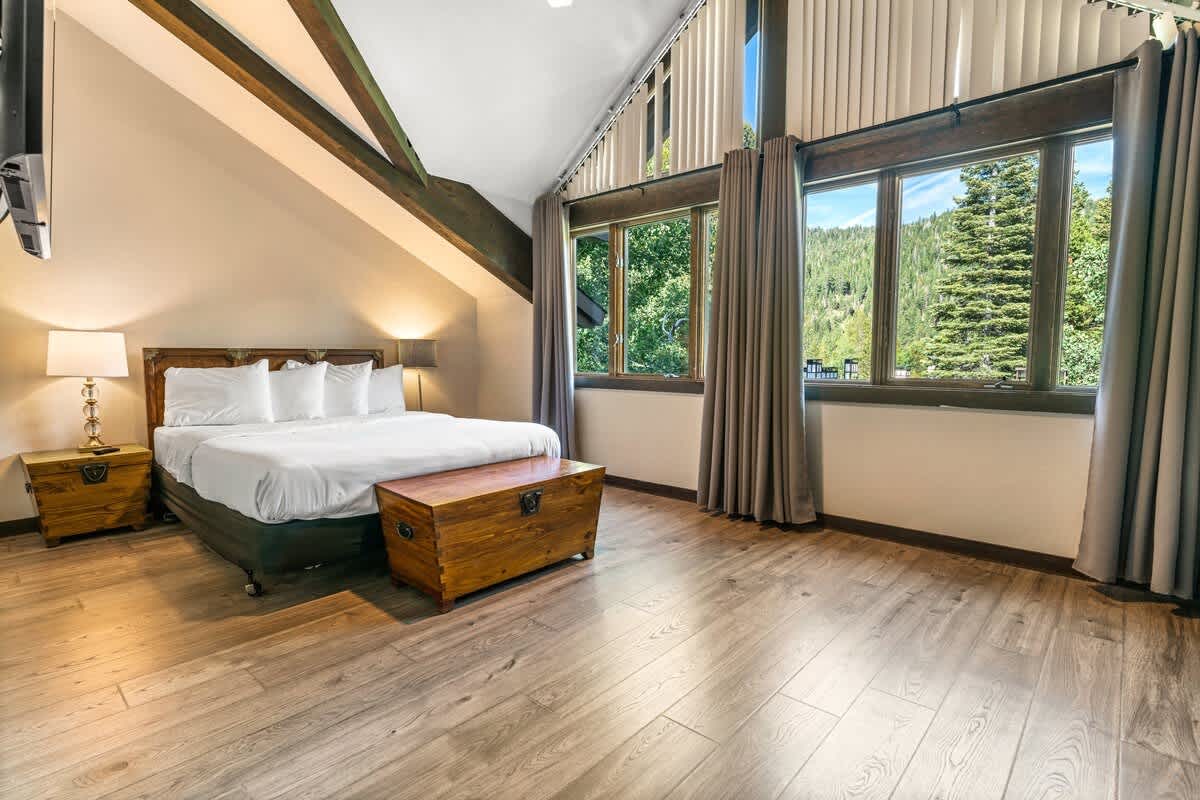 A cozy bedroom in an Olympic Valley vacation rental with a large bed, wooden furnishings, and forest views through tall windows.