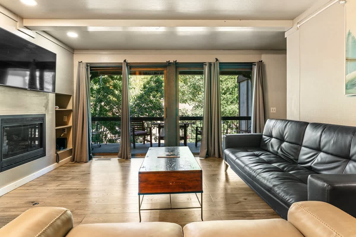 Modern living room in an Olympic Valley vacation rental, featuring a leather couch, large TV, and view of green trees.