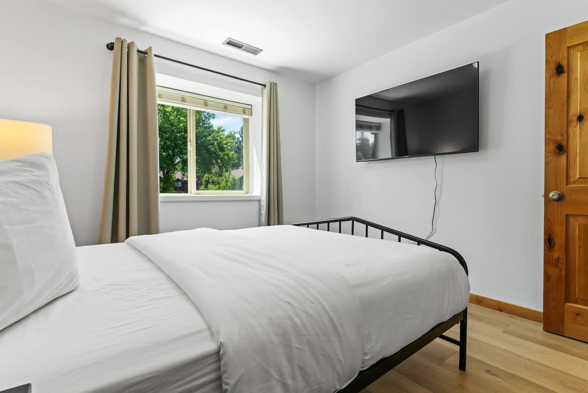 Bedroom in a Tahoe City vacation rental with a modern bed, wall-mounted TV, and a window view of greenery.