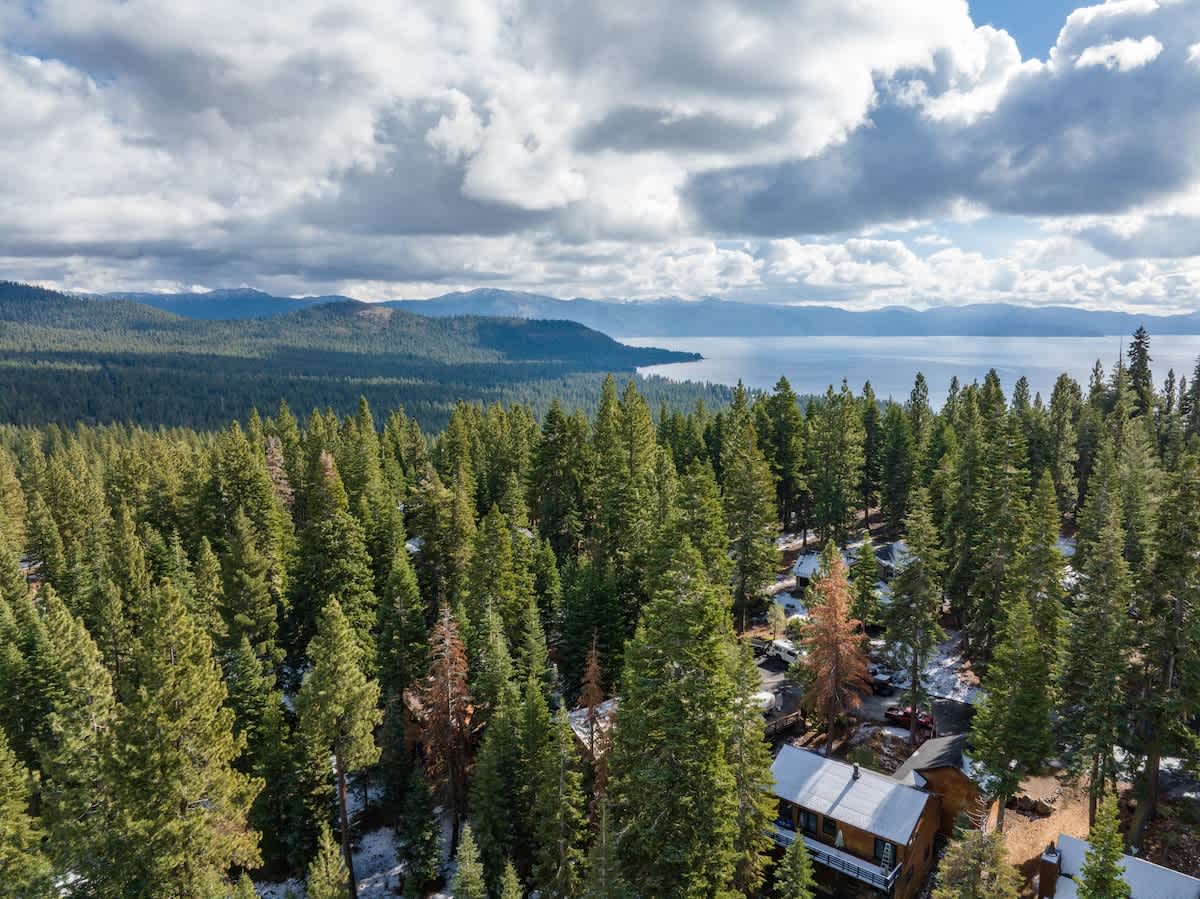 Aerial view of Tahoe Vista vacation rental surrounded by lush pine trees with a lake in the background under cloudy skies.