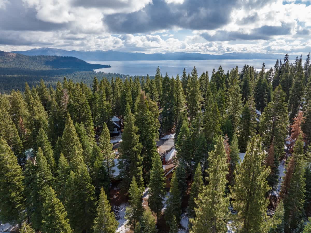 Aerial view of Tahoe Vista vacation rental surrounded by lush pine trees, overlooking a serene lake under cloudy skies.