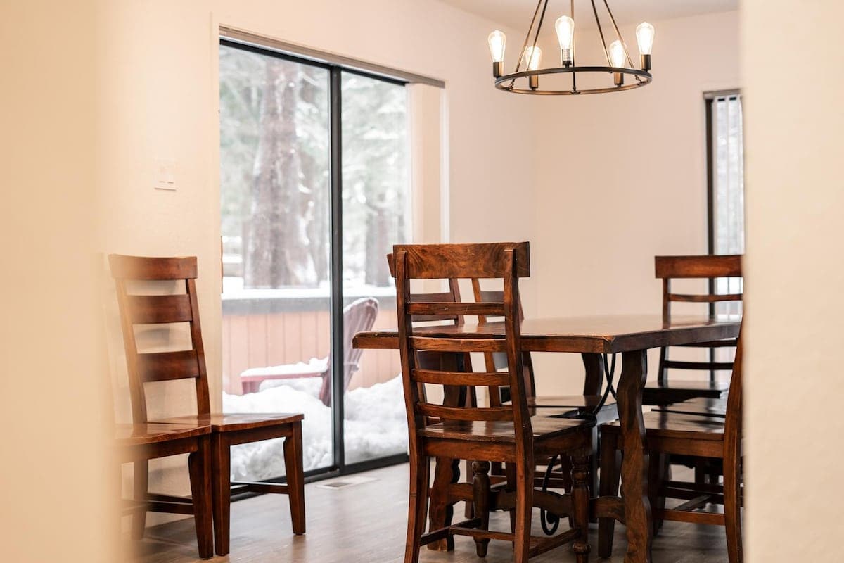 Cozy dining area in Truckee vacation rental with wooden table, chairs, and chandelier overlooking a snowy patio.