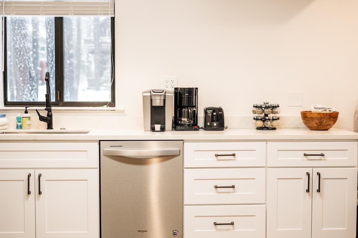 Modern kitchen in a Truckee vacation rental, featuring white cabinets, a coffee maker, and a view of snowy trees.