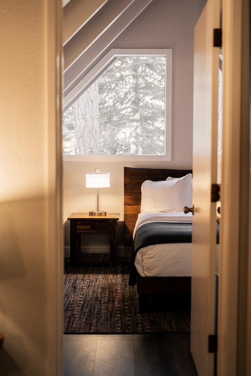 Cozy bedroom in a Tahoe Vista vacation rental, featuring a wooden bedframe, bedside lamp, and large window with forest view.