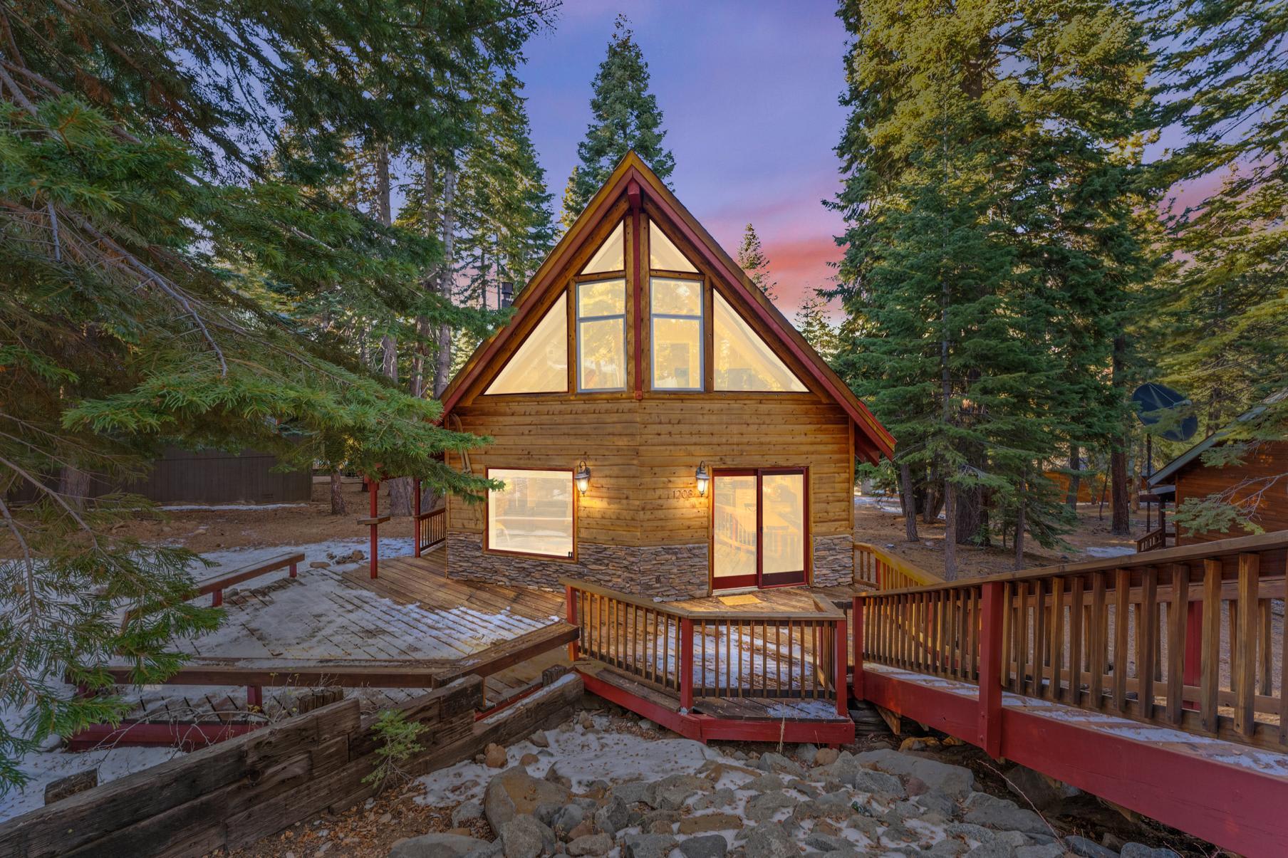 A-frame cabin vacation rental in Tahoe Vista surrounded by trees, with a wooden deck and large windows at sunset.