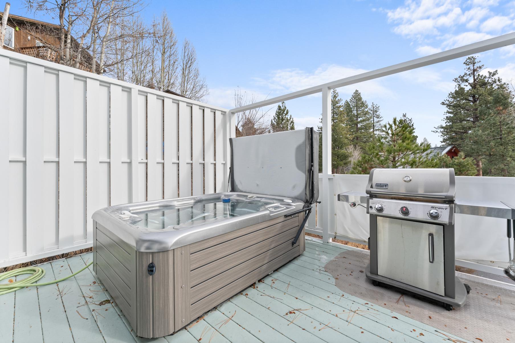 Deck of Truckee vacation rental with a hot tub and grill, surrounded by trees under a clear blue sky.