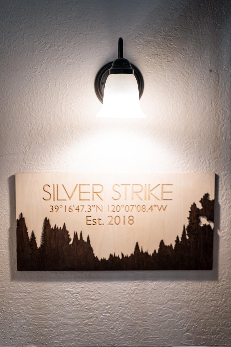 Wall-mounted sign reading "Silver Strike," coordinates, and "Est. 2018," illuminated by light in Truckee vacation rental.