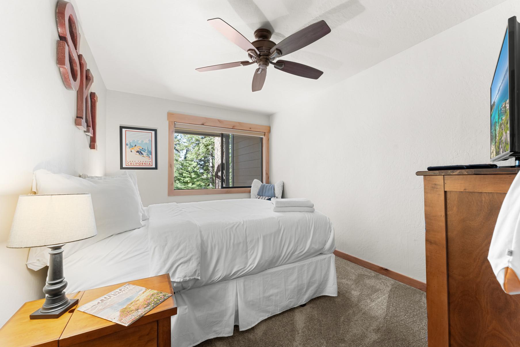 Cozy bedroom in a Truckee vacation rental with a large window, ceiling fan, and a TV on a wooden dresser.