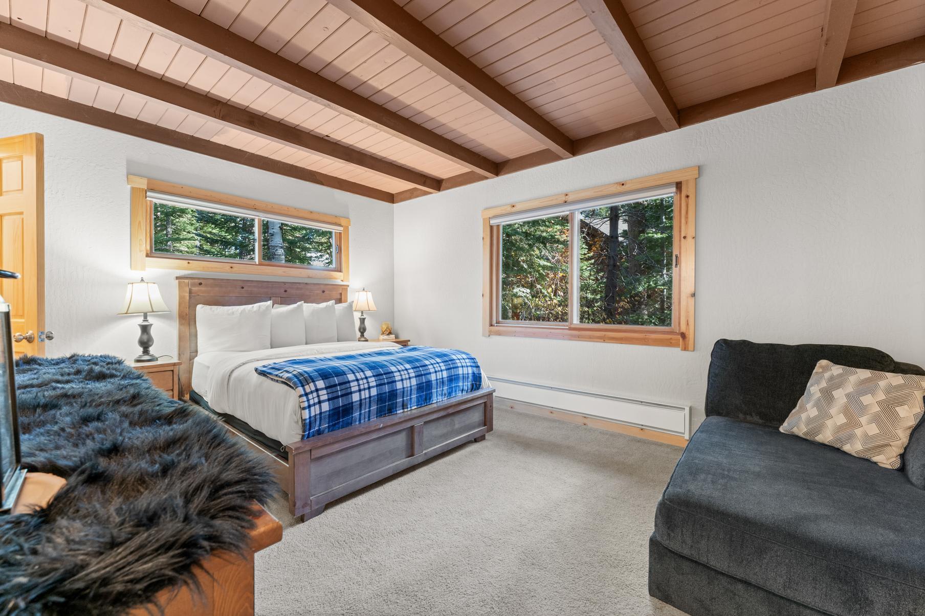 Cozy bedroom in Truckee vacation rental with a wooden bed, blue plaid blanket, large windows, and forest views.