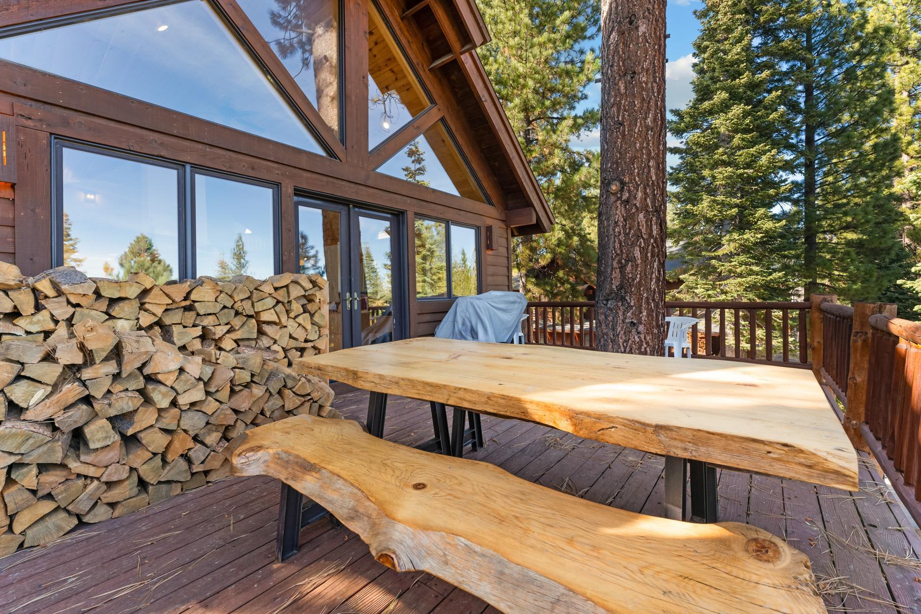 Rustic Truckee vacation rental patio with wooden tables, surrounded by tall pine trees and stacked firewood.