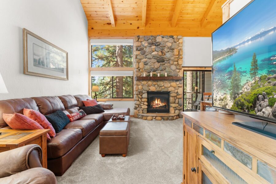 Cozy Truckee vacation rental with a leather sofa, stone fireplace, and large windows surrounded by trees.