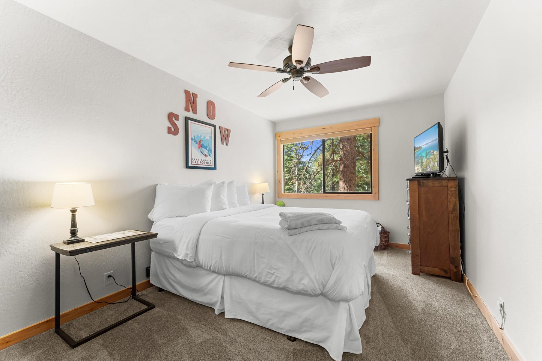 Cozy bedroom in Truckee vacation rental, features a plush bed, ceiling fan, and window view of forested landscape.