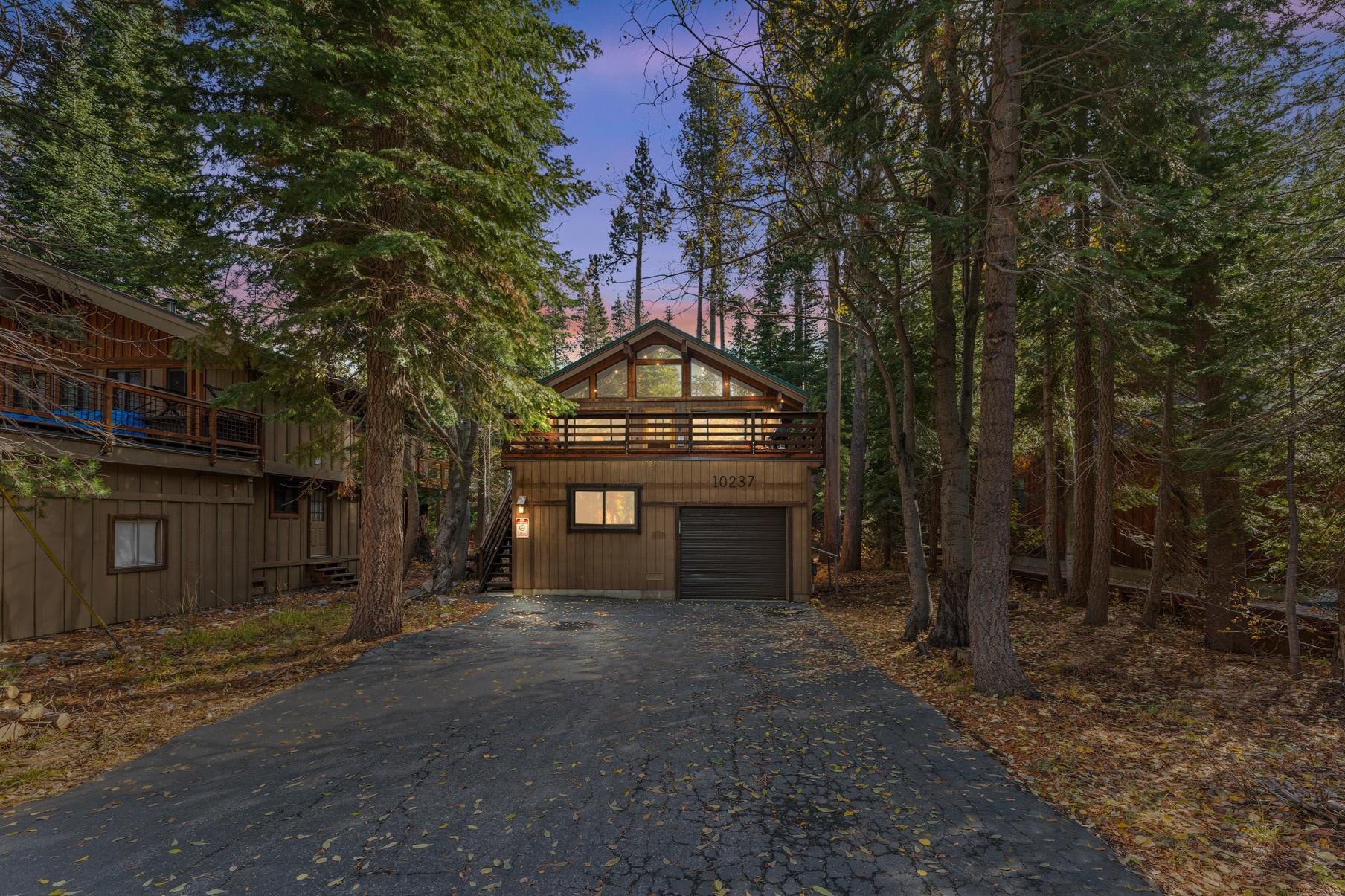 Cozy cabin-style vacation rental in Truckee, nestled among tall trees with a driveway leading to a garage and wooden deck.