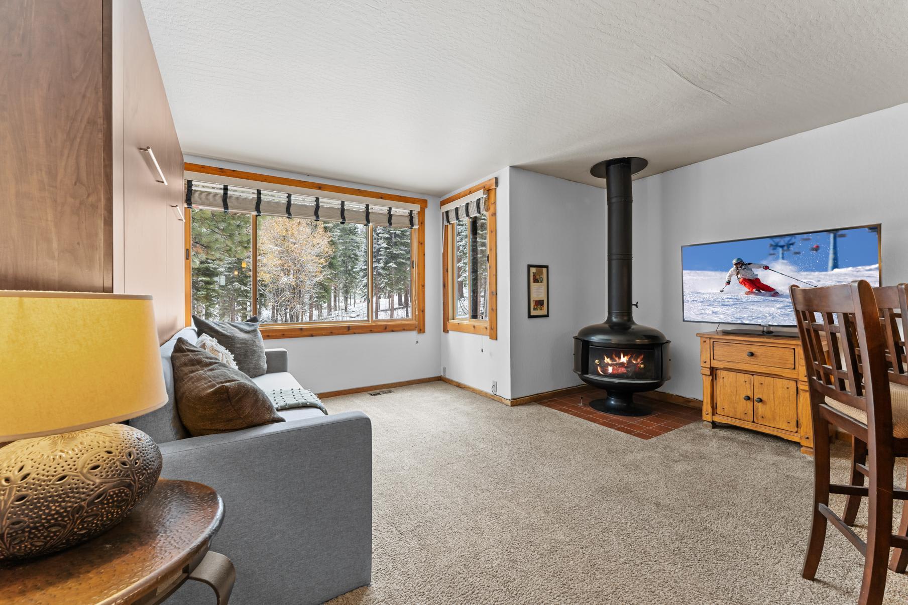 Cozy living room in a Truckee vacation rental with a view of snowy woods, a modern fireplace, and a TV showing skiing.