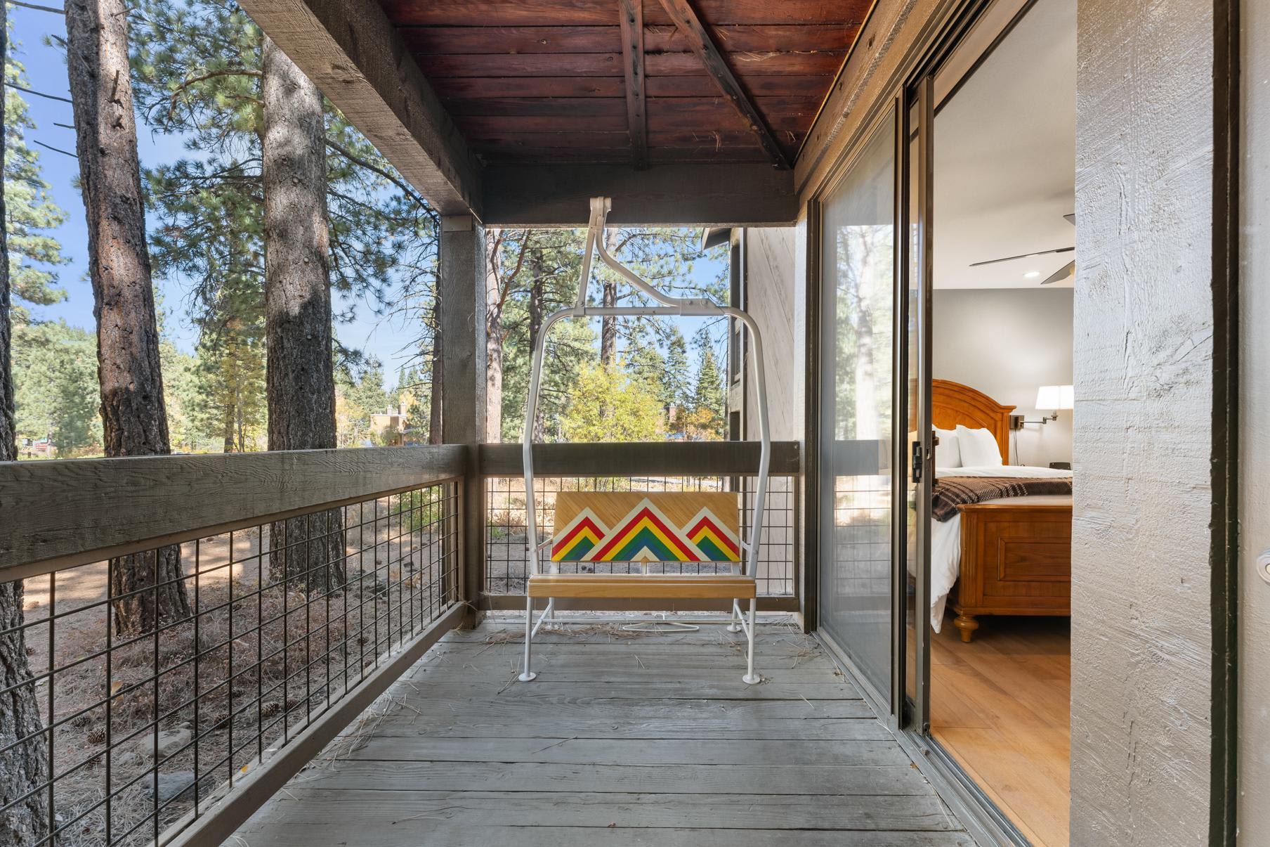 Covered balcony with a colorful chair, overlooking pine trees at a Truckee vacation rental, next to a cozy bedroom.