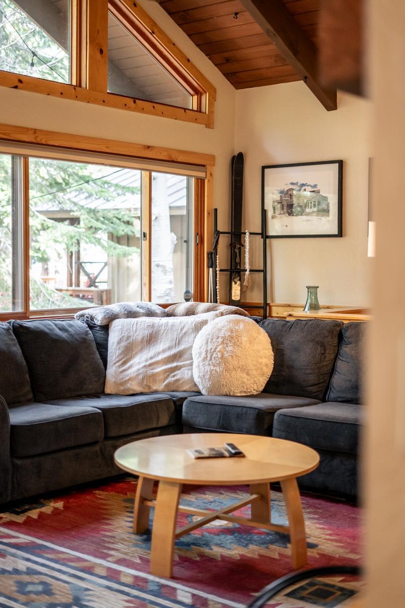 Cozy Truckee vacation rental with a plush gray sofa, wooden table, large windows, and a colorful rug.