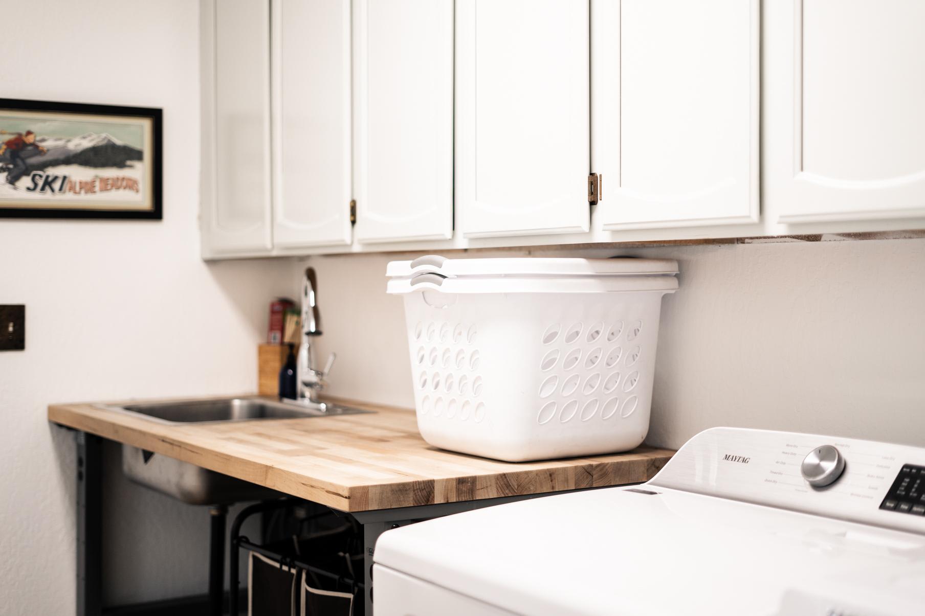 Laundry room in Truckee vacation rental with basket on wooden counter, washer, and retro ski poster on wall.