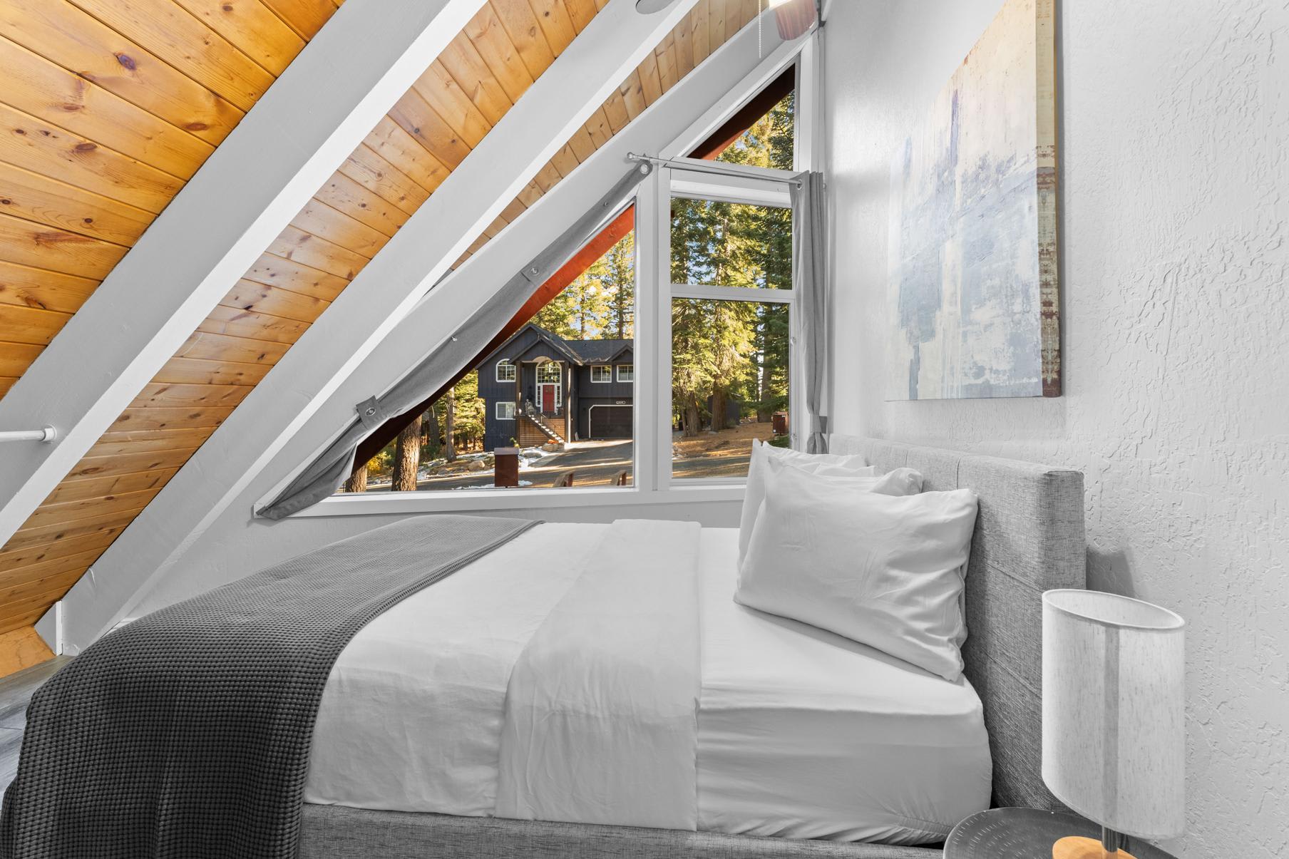 A-frame bedroom in a Tahoe Vista vacation rental with a cozy bed, wooden ceiling, and window view of trees and chalet.