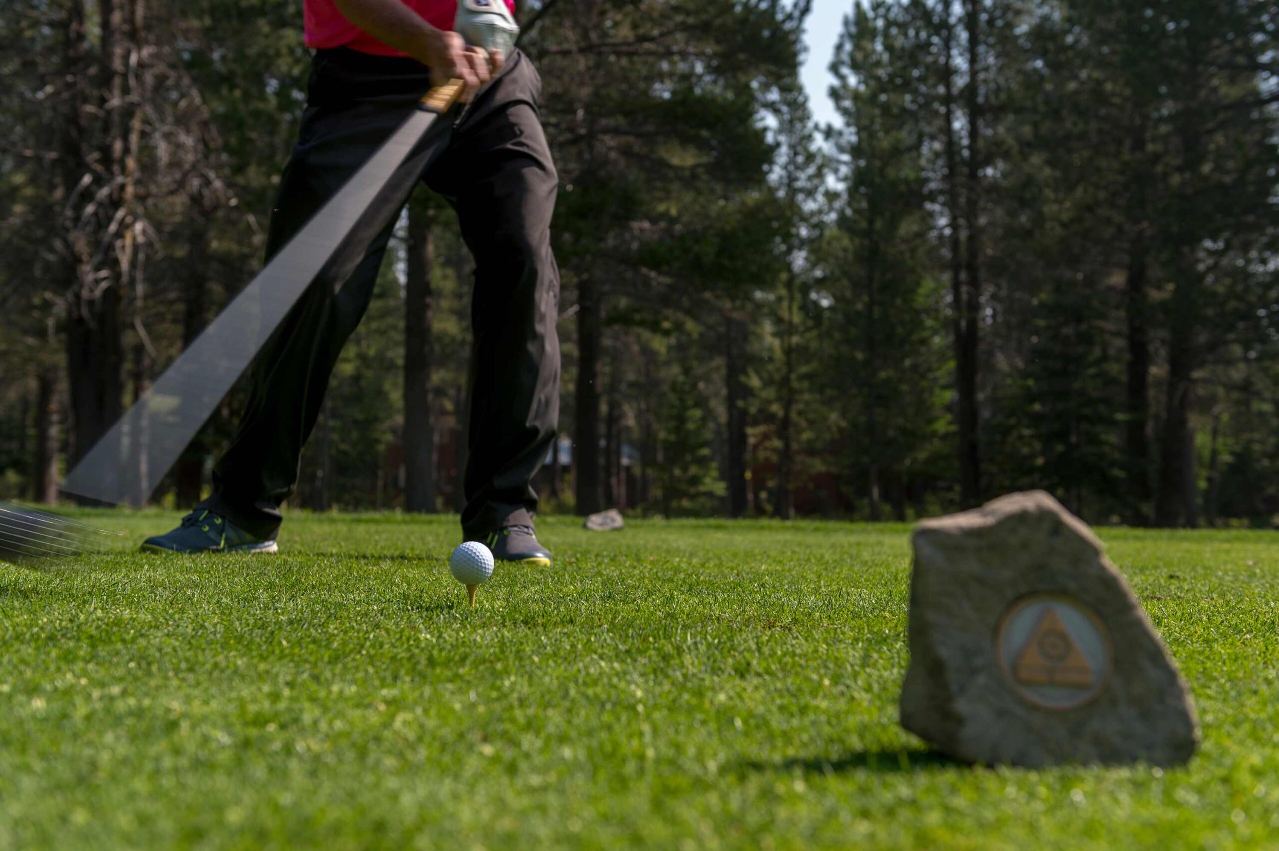 Golfer swings at a ball on a green course near a rock marker, set in a wooded area in Truckee, ideal for a vacation rental.