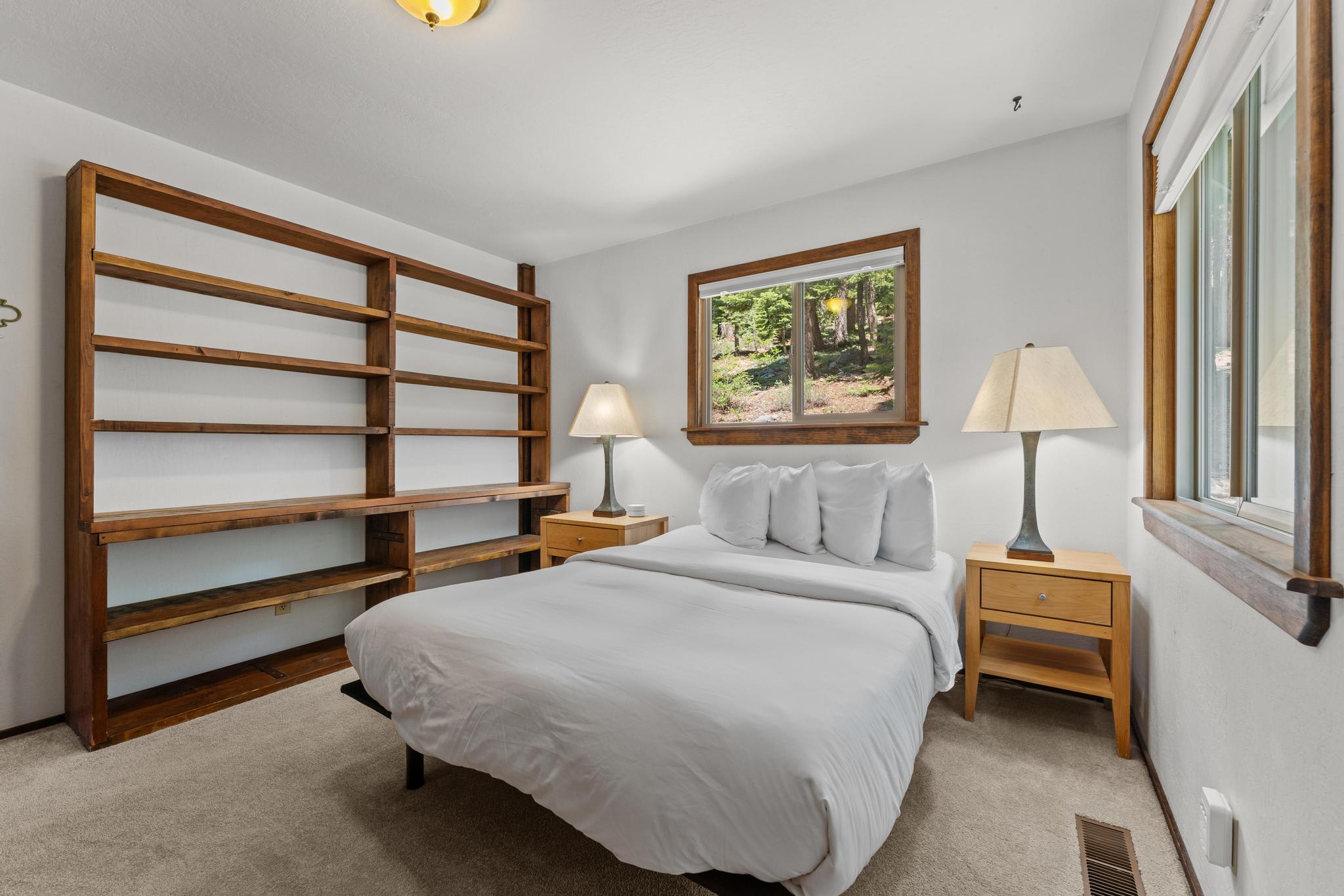 Bedroom in Tahoe Vista vacation rental with a cozy bed, wooden shelves, nightstands, lamps, and a forest view window.