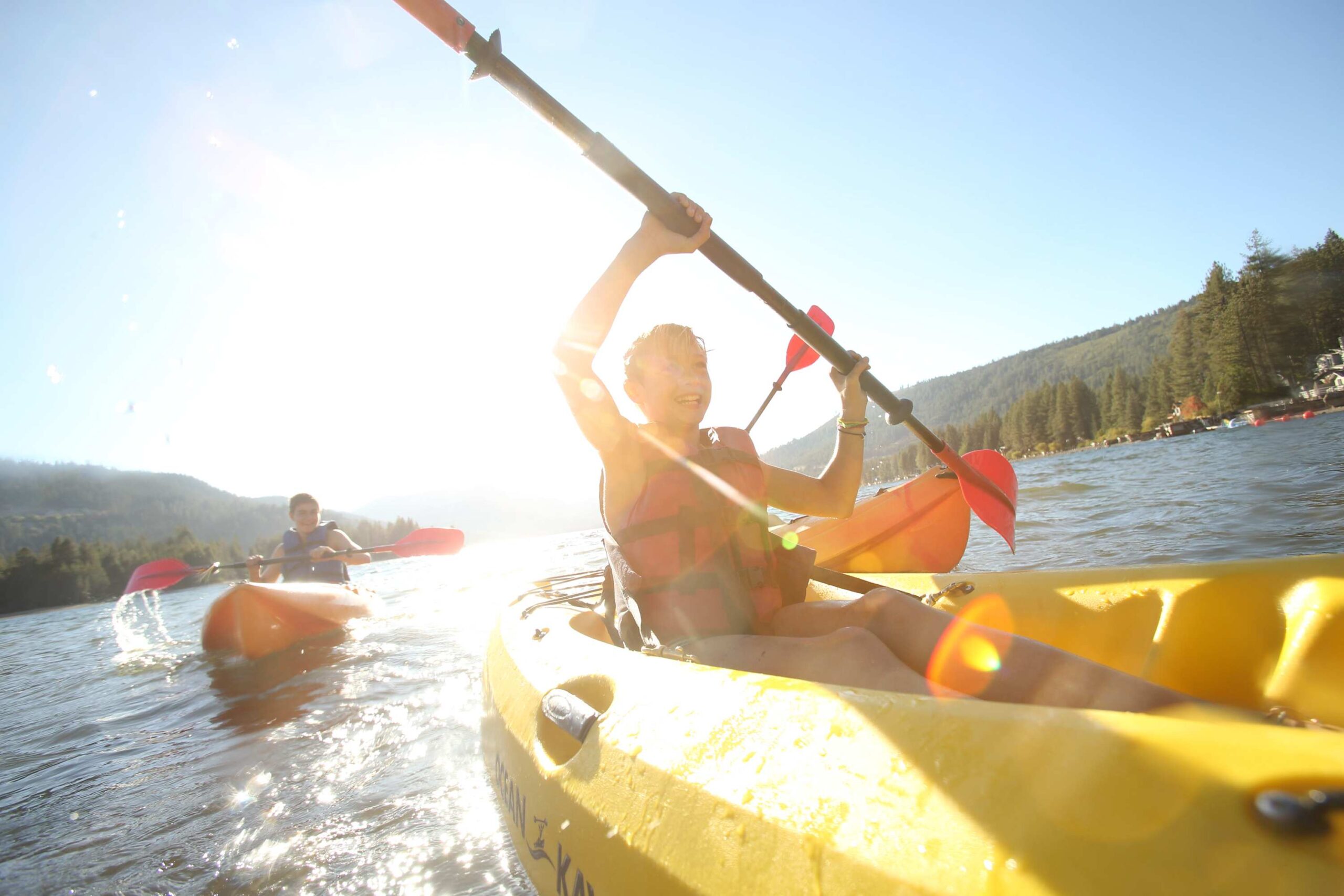 Two people kayaking on a sunny lake in Truckee, near a vacation rental, surrounded by forested hills.