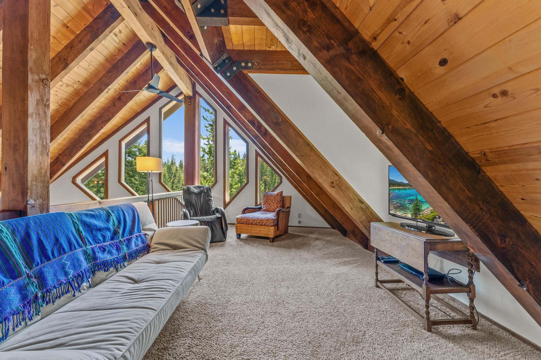 Rustic Tahoe Vista vacation rental loft with wooden beams, cozy seating, and forest views through triangular windows.
