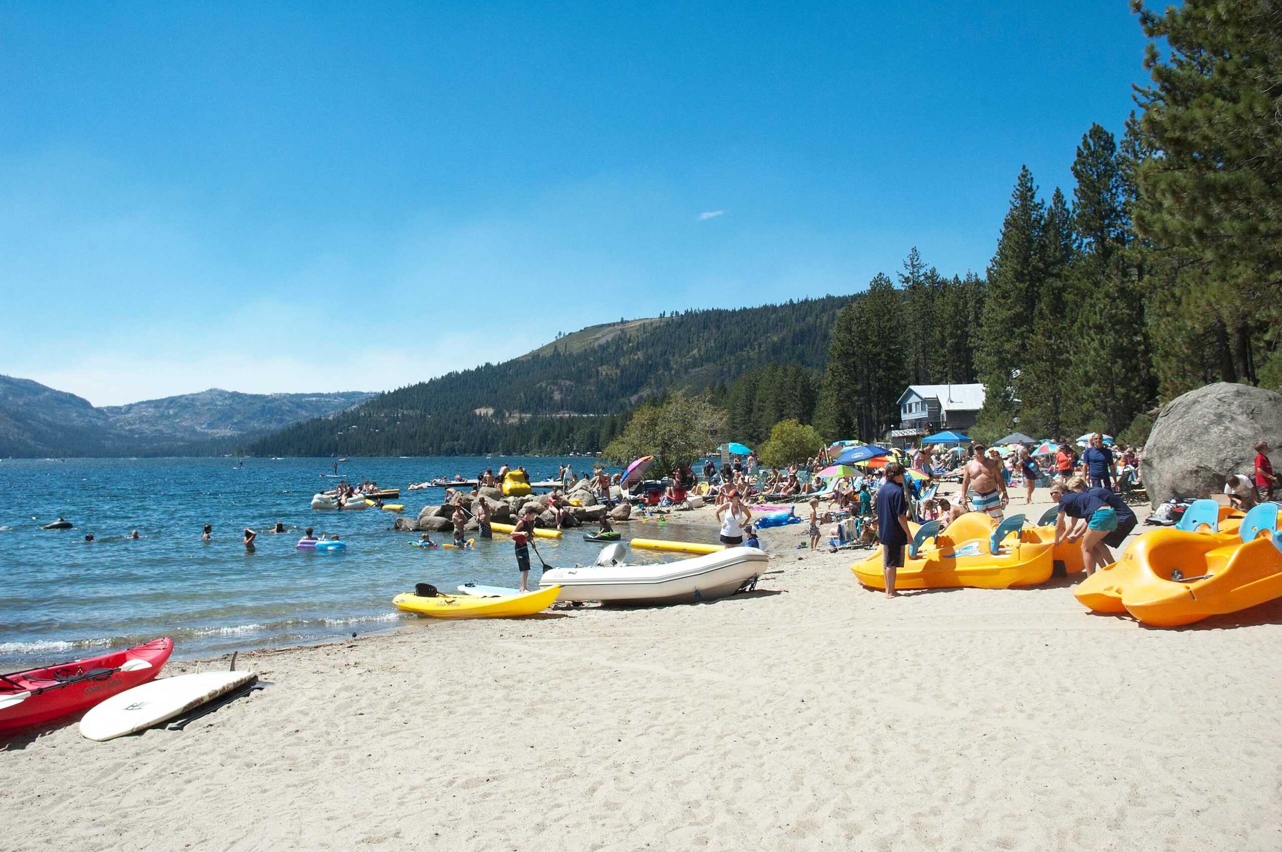 Beachgoers enjoy water activities near a vacation rental in Truckee, surrounded by mountains and trees under a blue sky.