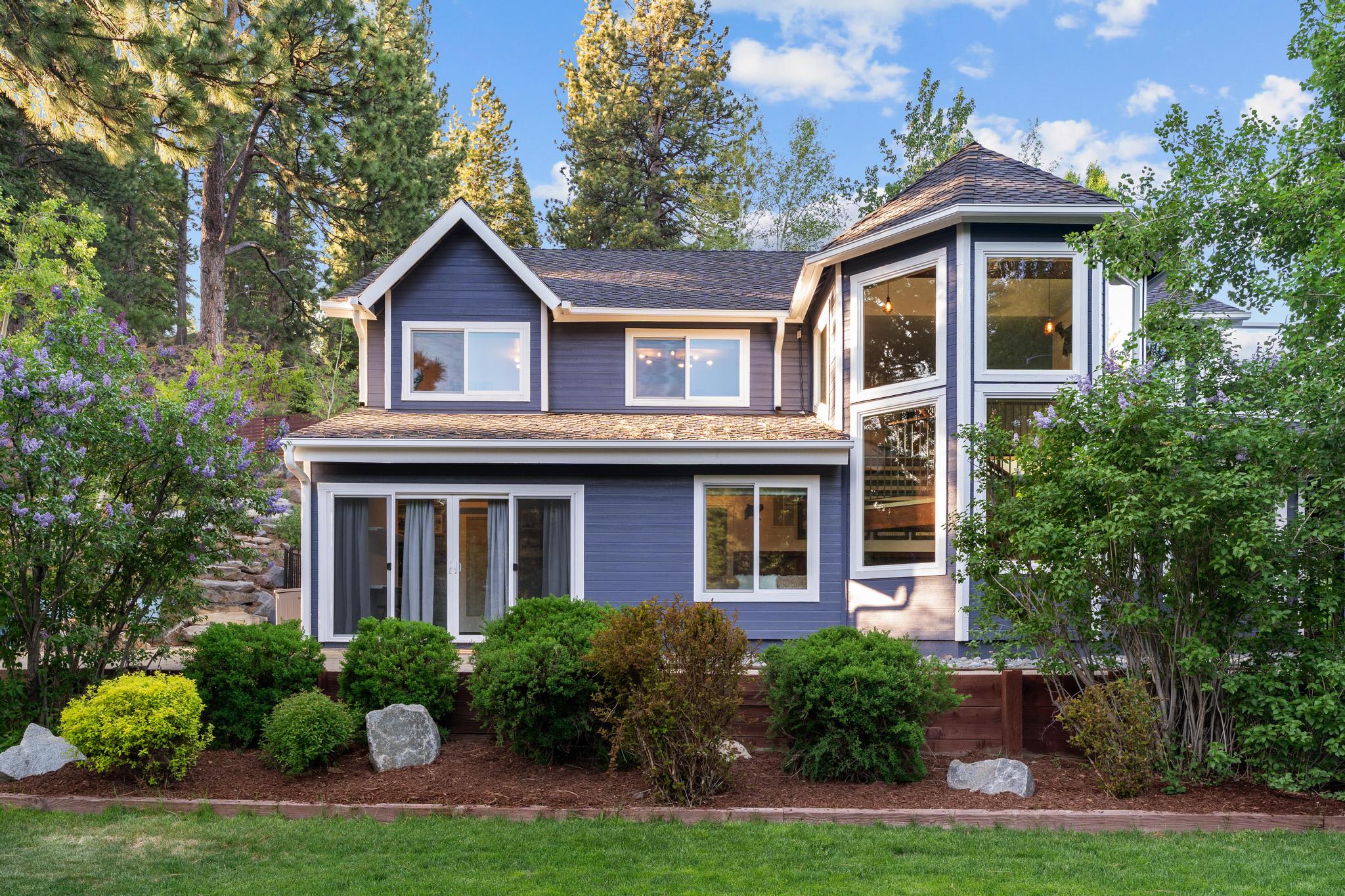 Charming Truckee vacation rental with blue exterior, lush greenery, and large windows surrounded by tall pine trees.