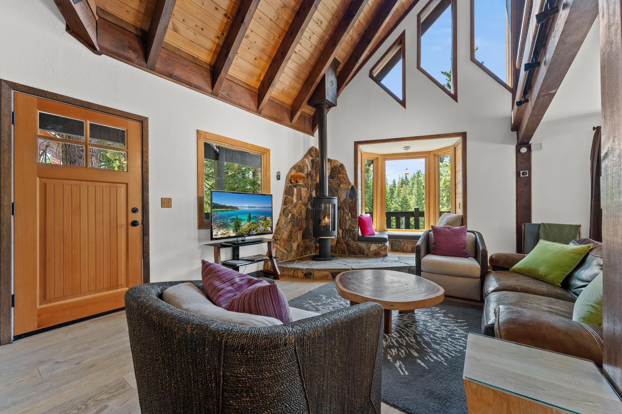 Cozy Tahoe Vista vacation rental with wooden beams, stone fireplace, leather sofa, and forest views through large windows.