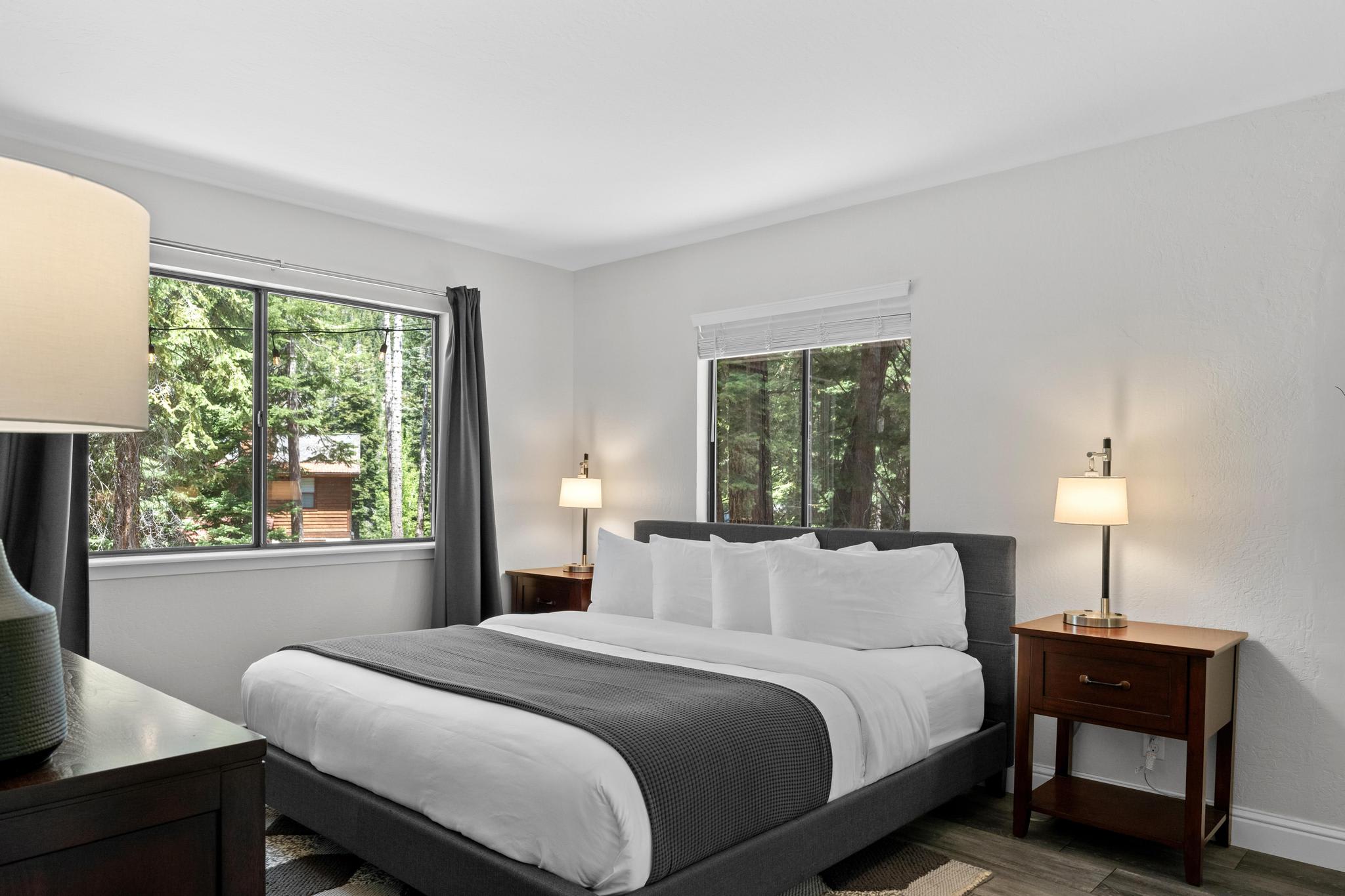 Cozy bedroom in a Tahoe Vista vacation rental, featuring a plush bed, nightstands, and large windows showing forest views.