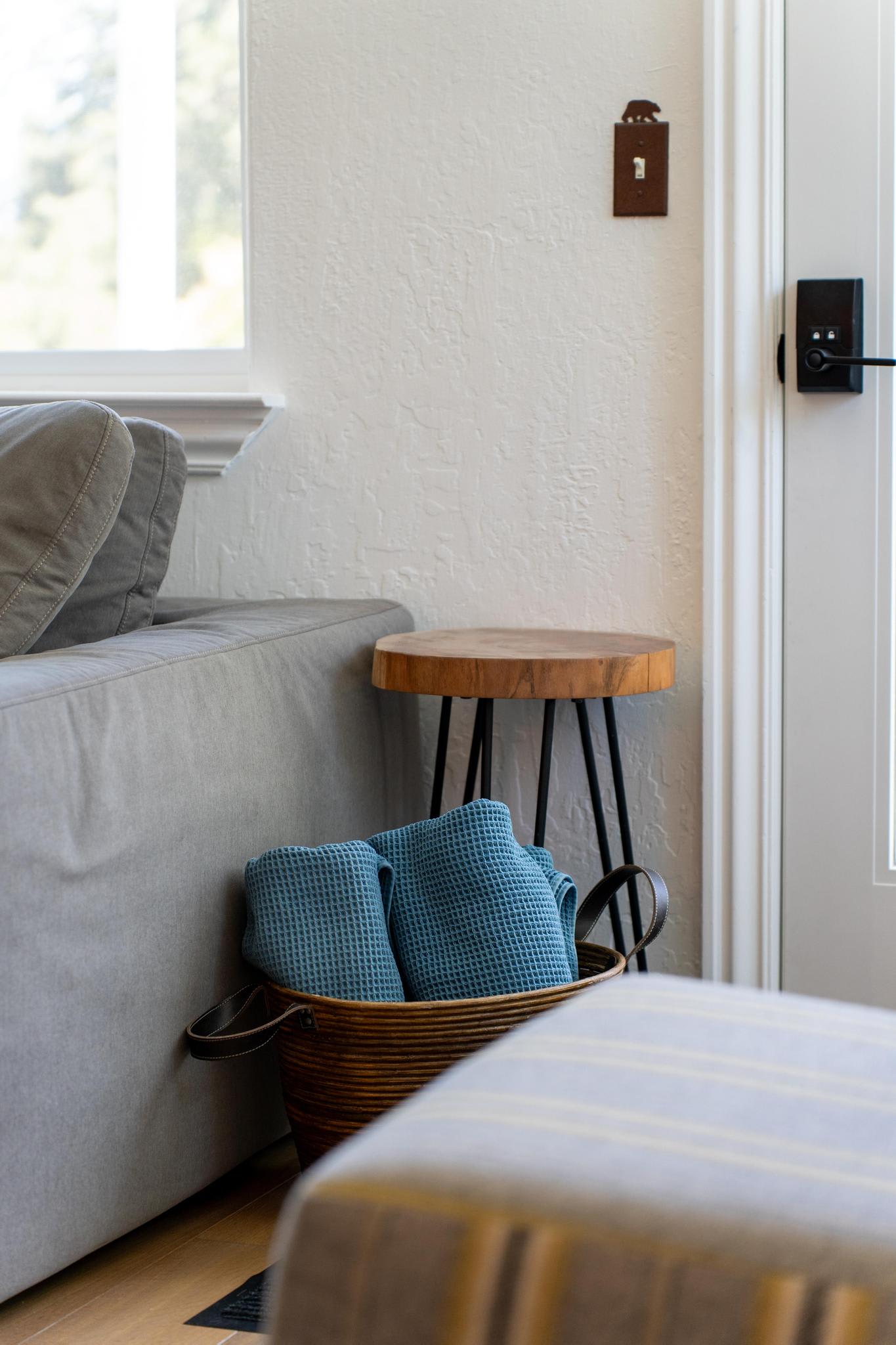 Cozy Truckee vacation rental with a gray sofa, wooden side table, and basket of blue towels by a door with natural light.