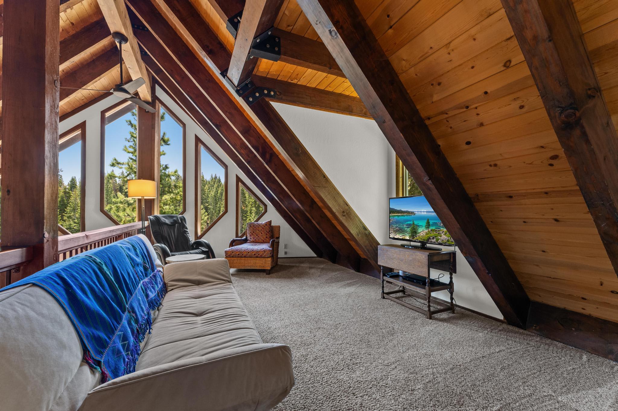 Loft in Tahoe Vista vacation rental with rustic wooden beams, cozy seating, TV, and forest views through triangular windows.