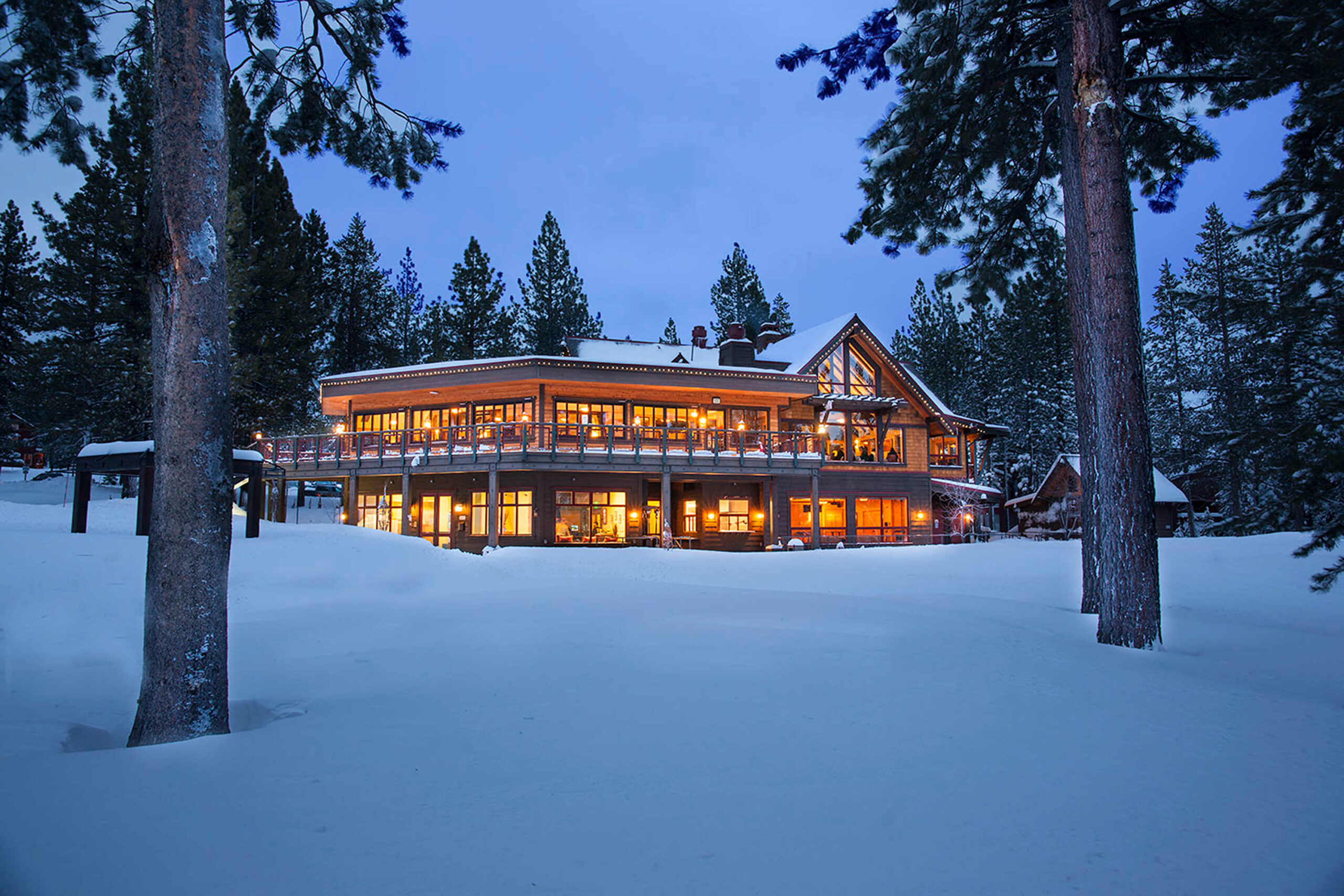 Cozy, illuminated vacation rental in snowy Truckee, surrounded by tall trees against a deep blue evening sky.