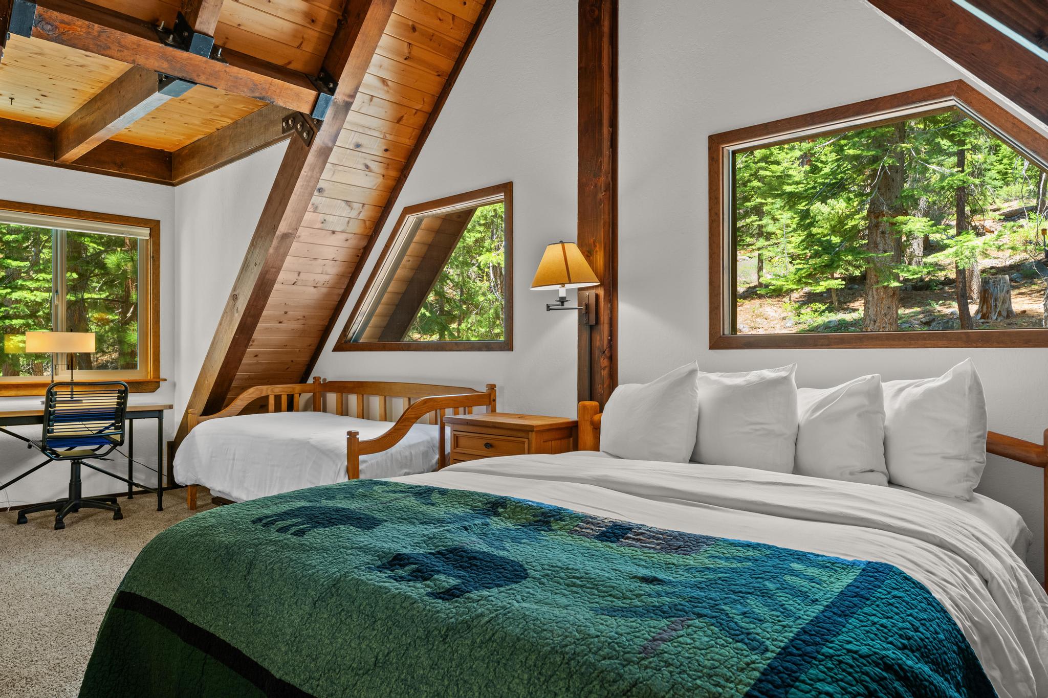 Cozy bedroom in Tahoe Vista vacation rental with wooden beams, green quilt, and views of lush trees through large windows.
