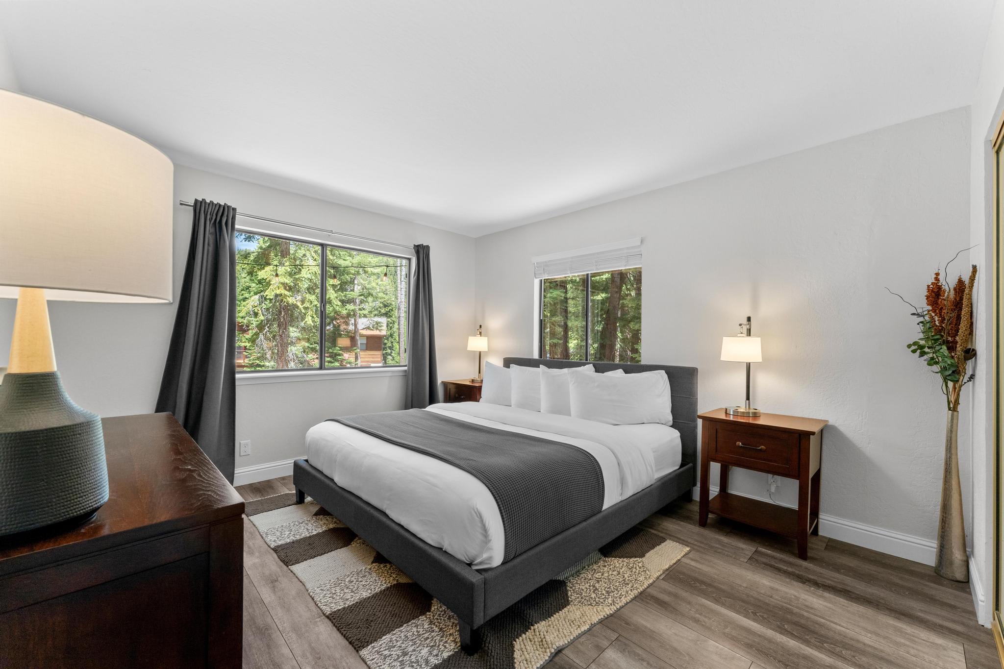 Modern bedroom in Tahoe Vista vacation rental with large windows, a cozy bed, lamps, and forest views.