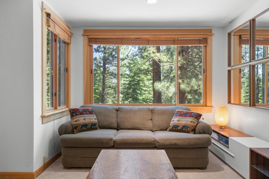 Cozy Truckee vacation rental with sofa, bear-themed pillows, large windows offering a view of lush pine trees.