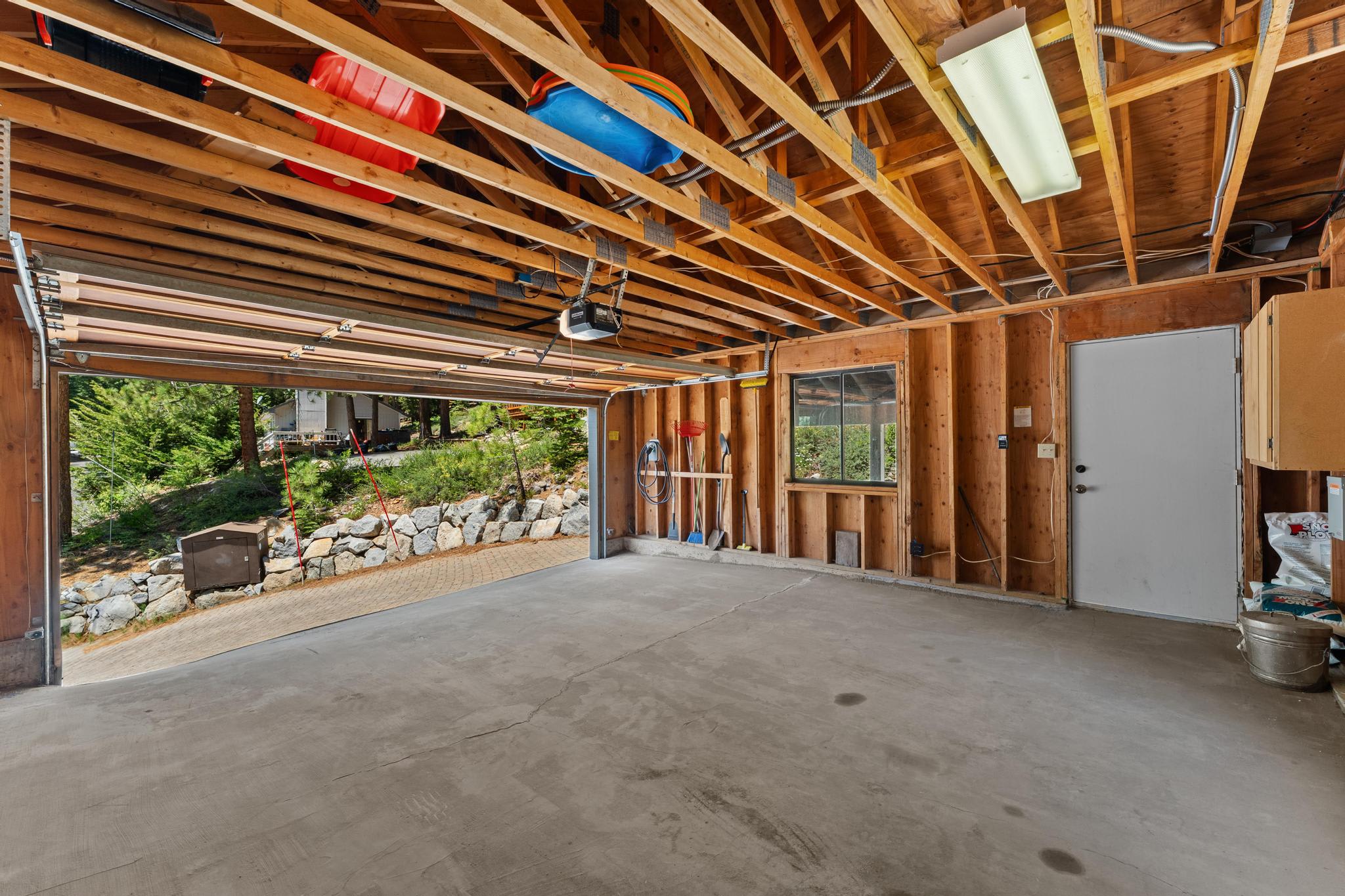 Open garage with concrete floor and wooden walls in a Tahoe Vista vacation rental. Exterior view shows greenery and stone wall.