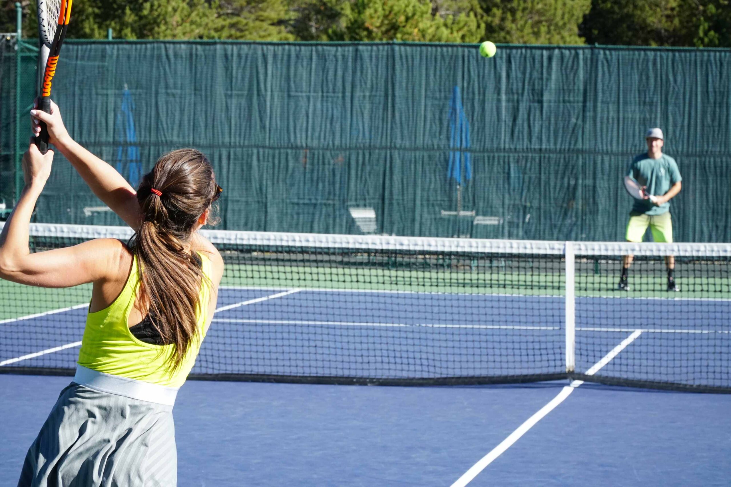 A woman and man play tennis at a Truckee vacation rental, surrounded by trees and a green fence.