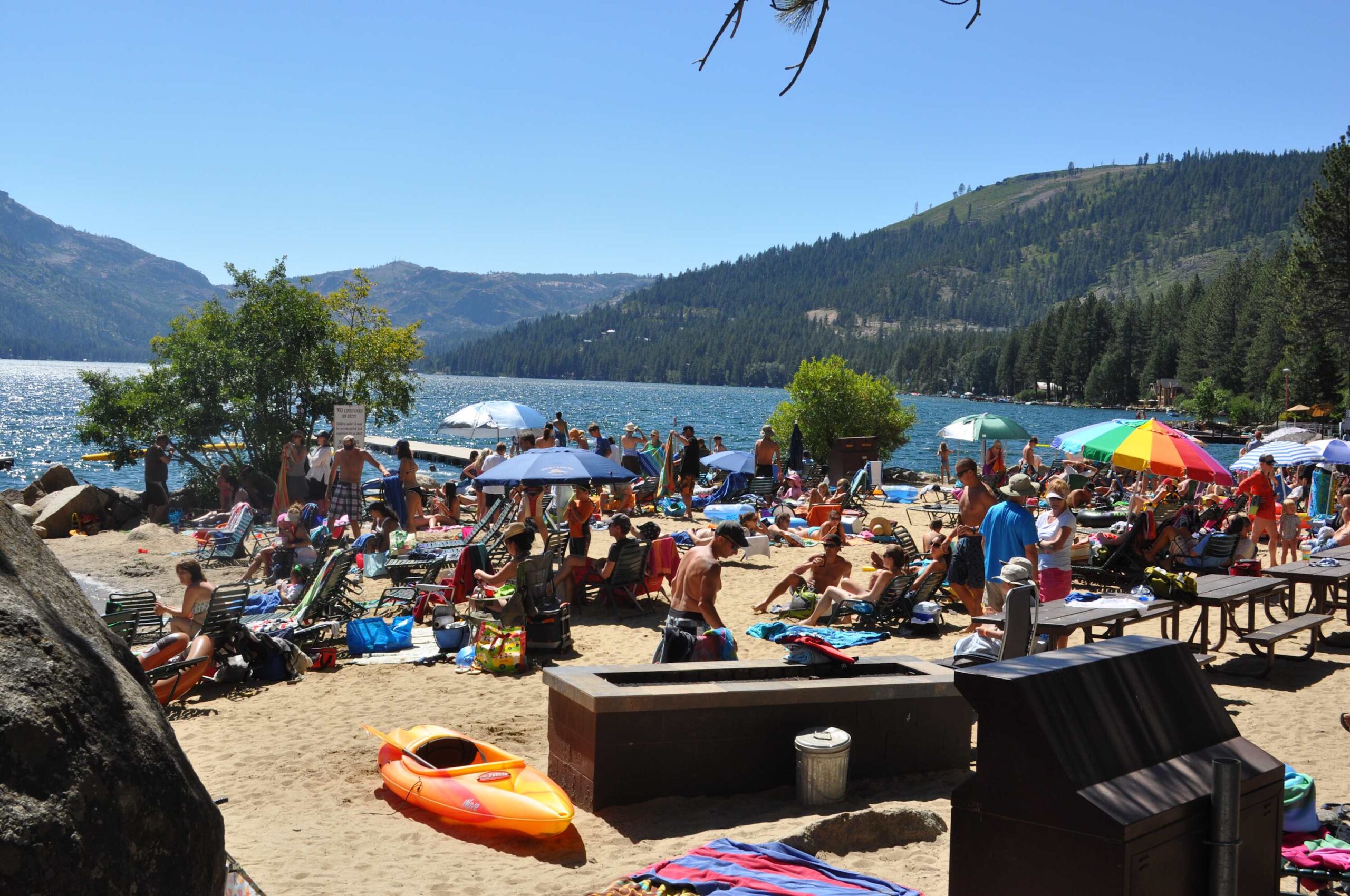 Crowded sandy beach by a lake in Truckee, with umbrellas and kayaks. Ideal spot for a vacation rental getaway.