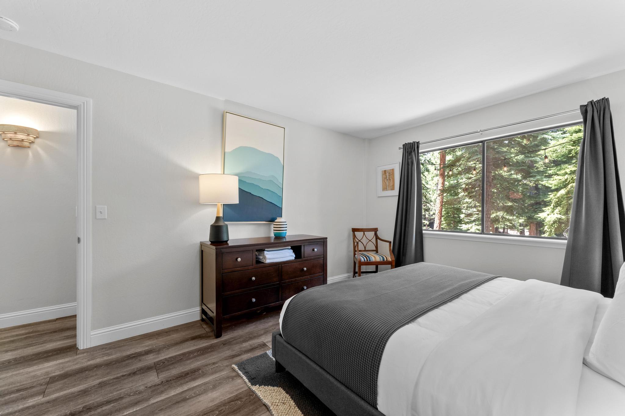 Bedroom in Tahoe Vista vacation rental with wooded view, cozy bed, dresser, and lamp on wooden floor.