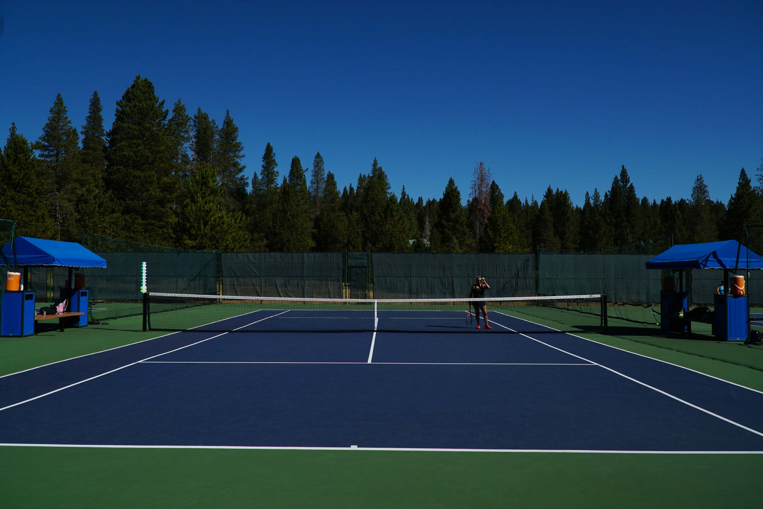 Tennis court surrounded by forest at a vacation rental in Truckee, with a person playing under a clear blue sky.