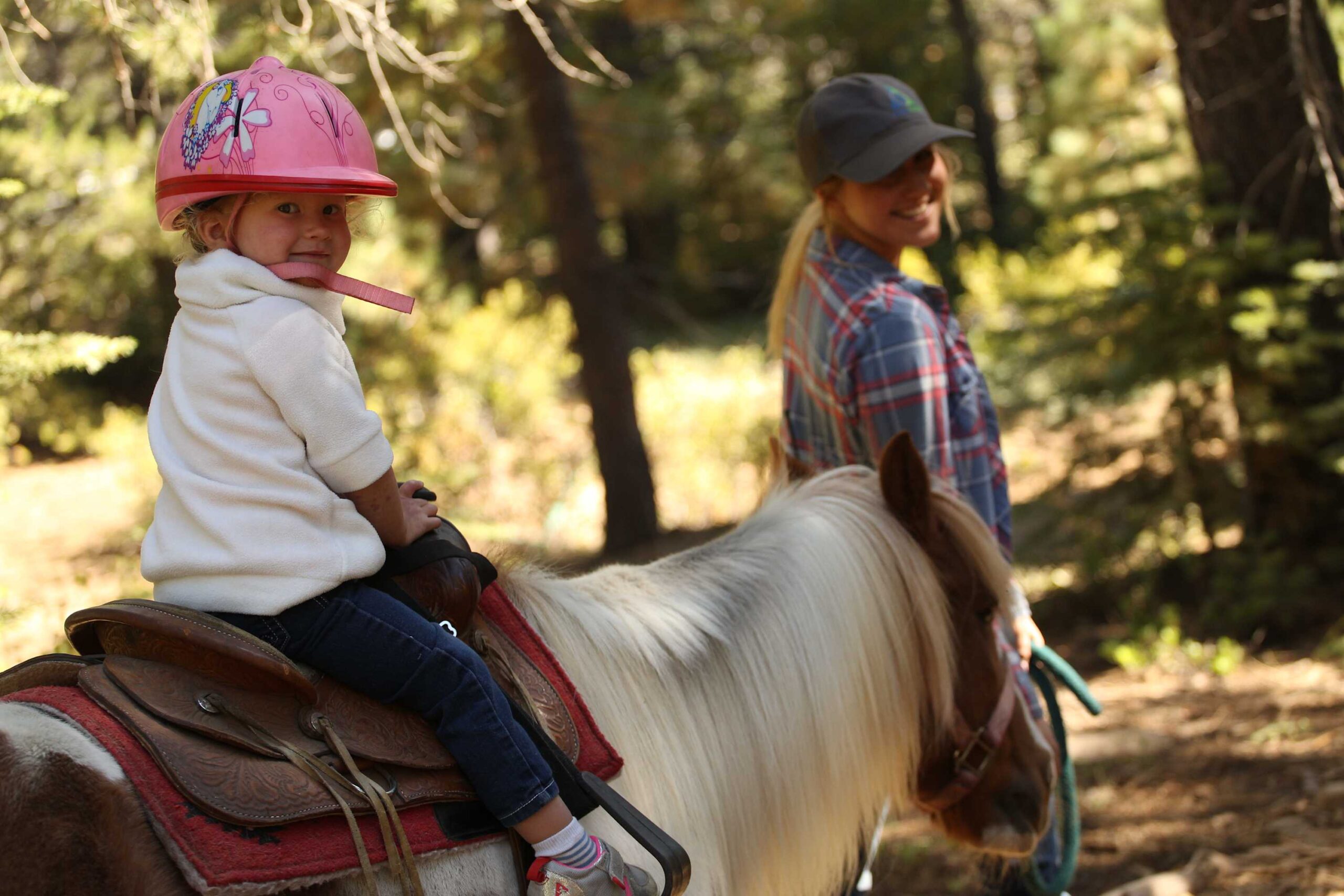 Child riding a pony in a forest near Truckee, led by a woman, experiencing a vacation rental activity.