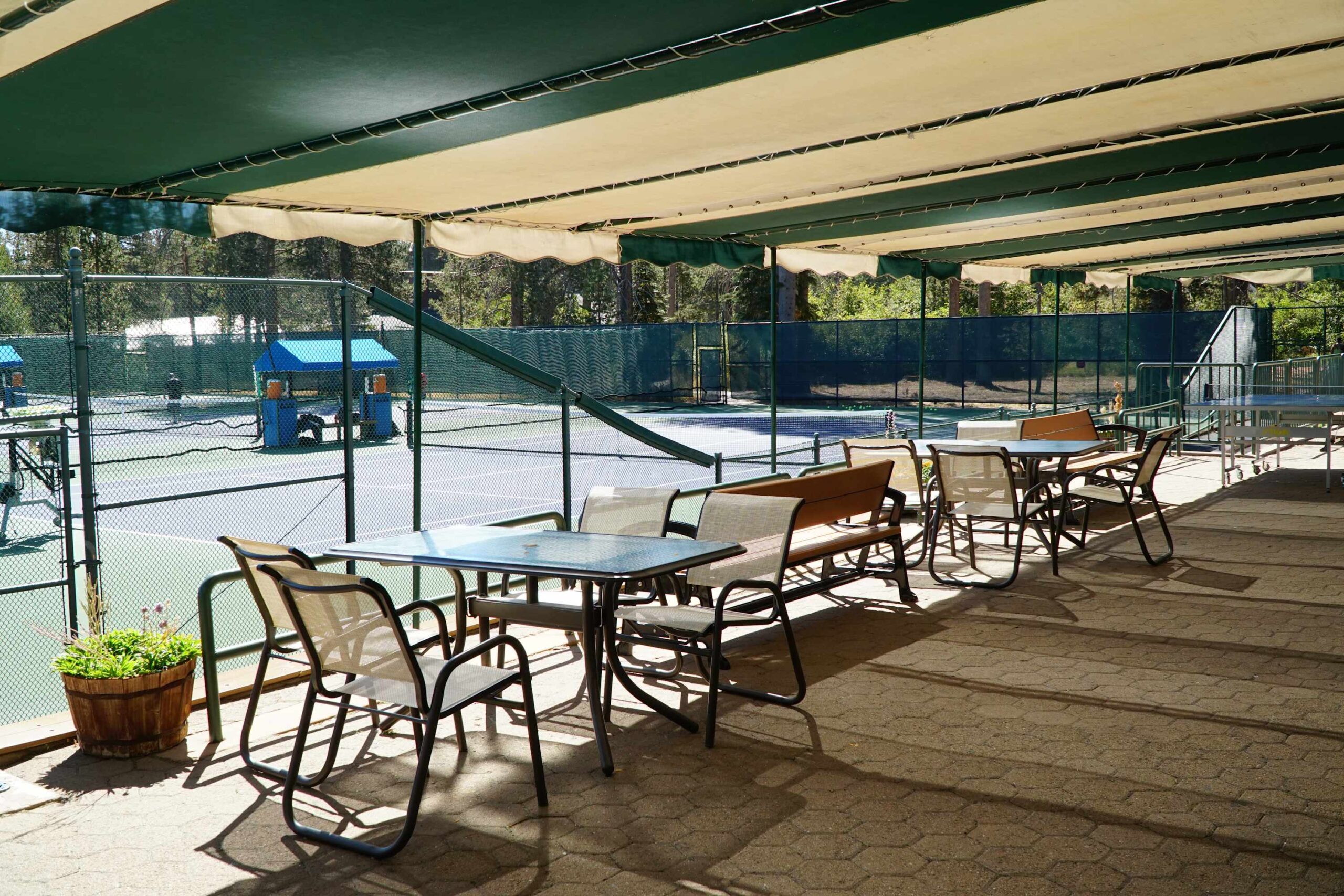 Shaded patio with tables overlooking a tennis court at a Truckee vacation rental, surrounded by trees.