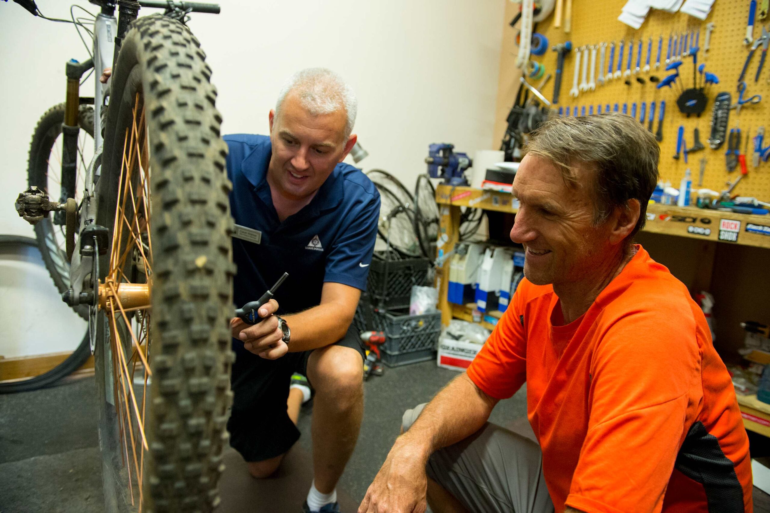 Two men in a workshop fixing a bike in Truckee near a vacation rental, surrounded by tools and equipment.