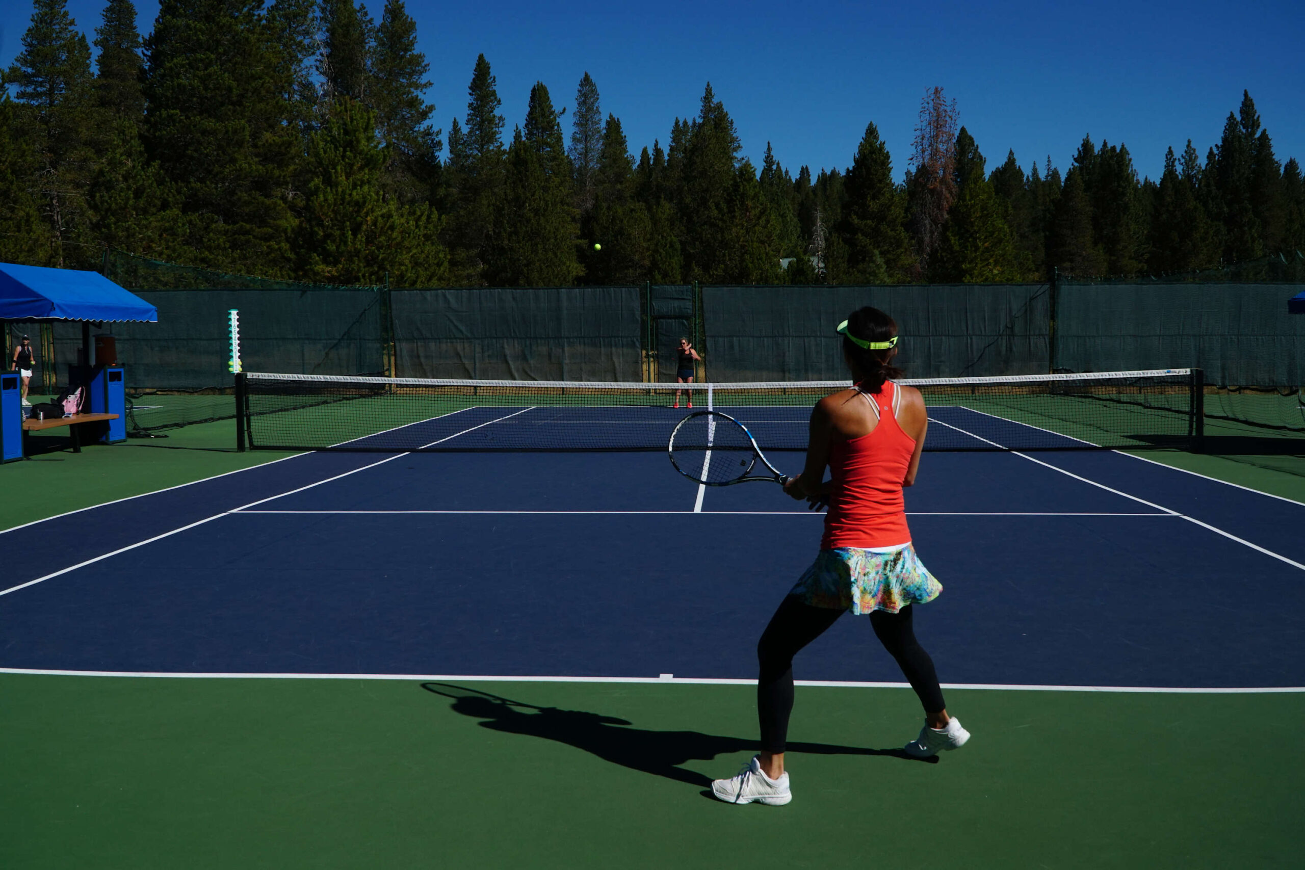 Two people playing tennis at a vacation rental in Truckee, surrounded by lush pine trees under a clear blue sky.