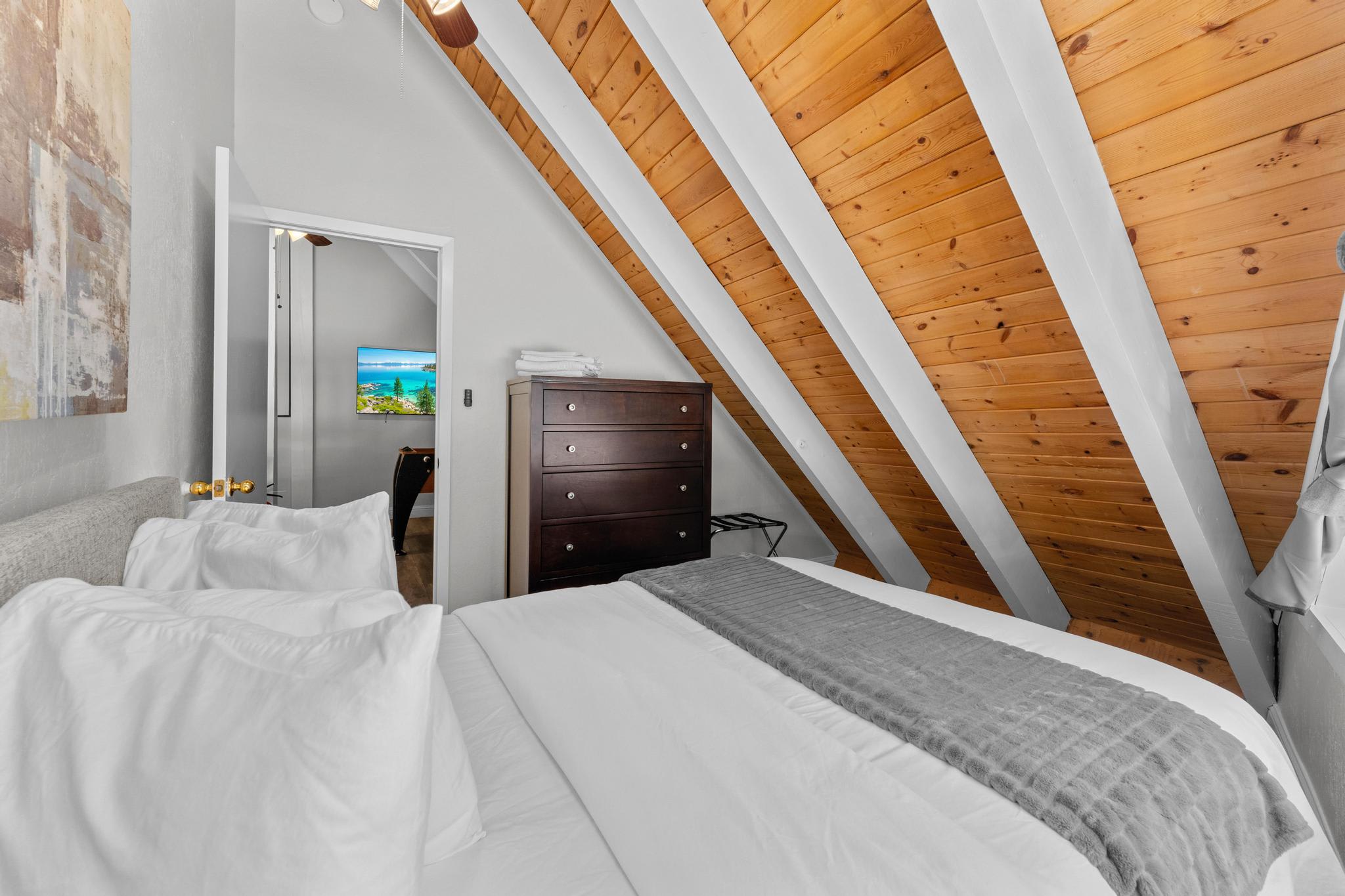 A cozy Tahoe Vista vacation rental bedroom with slanted wooden ceiling, white bedding, dresser, and a wall-mounted TV.