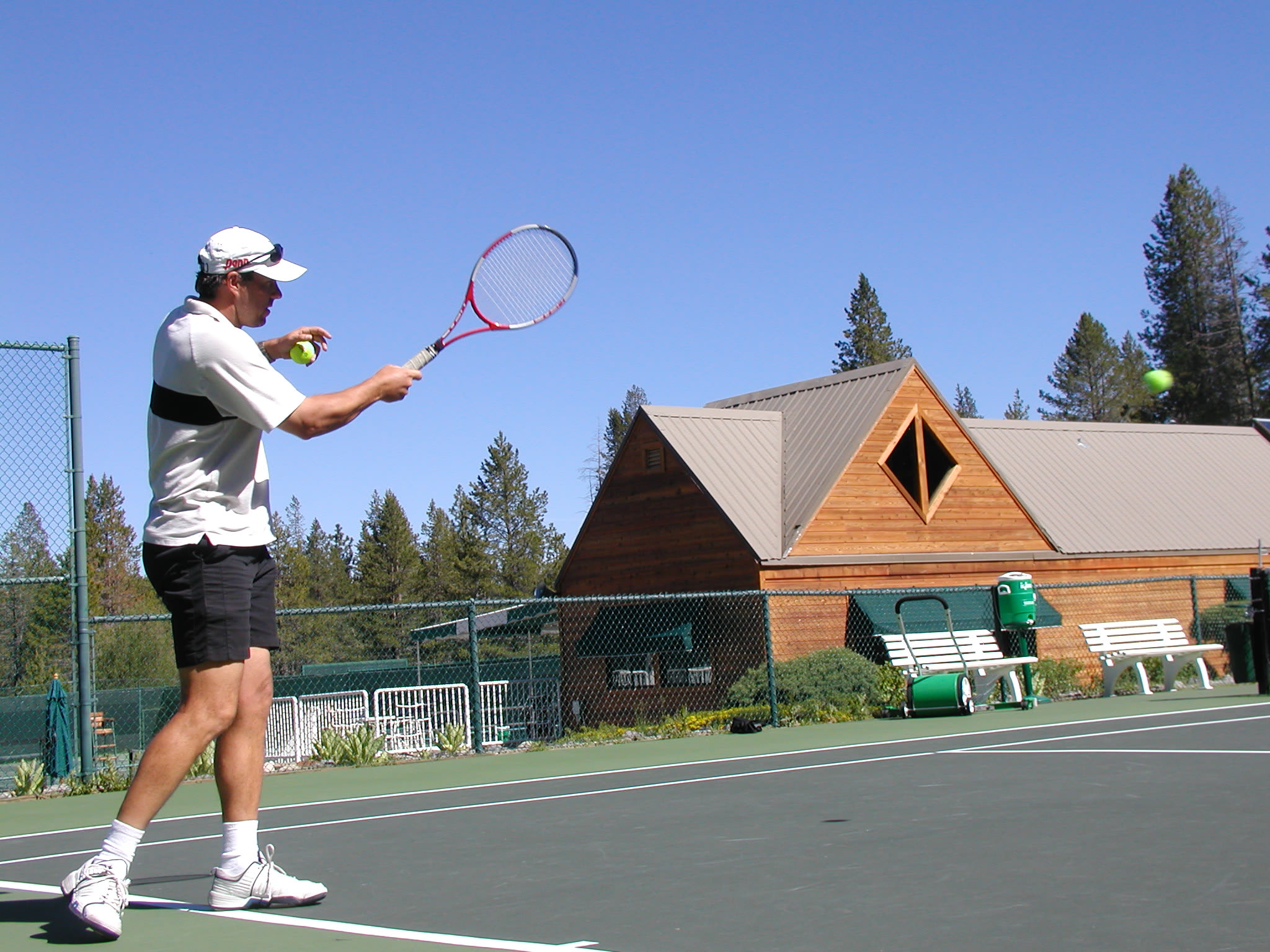 Man playing tennis at a court near a rustic vacation rental in Truckee, surrounded by pine trees under a clear blue sky.