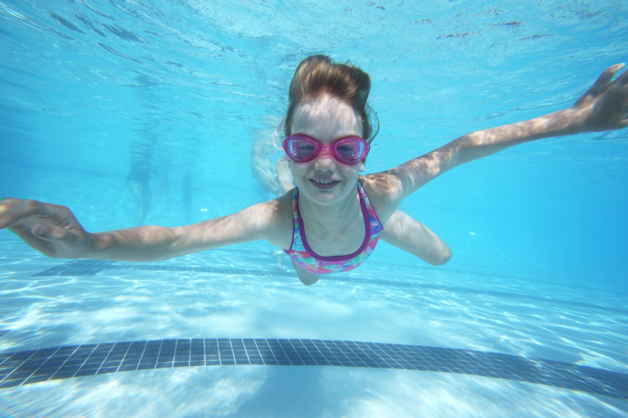 Young girl swimming underwater in a pool at a Truckee vacation rental, wearing pink goggles and a colorful swimsuit.