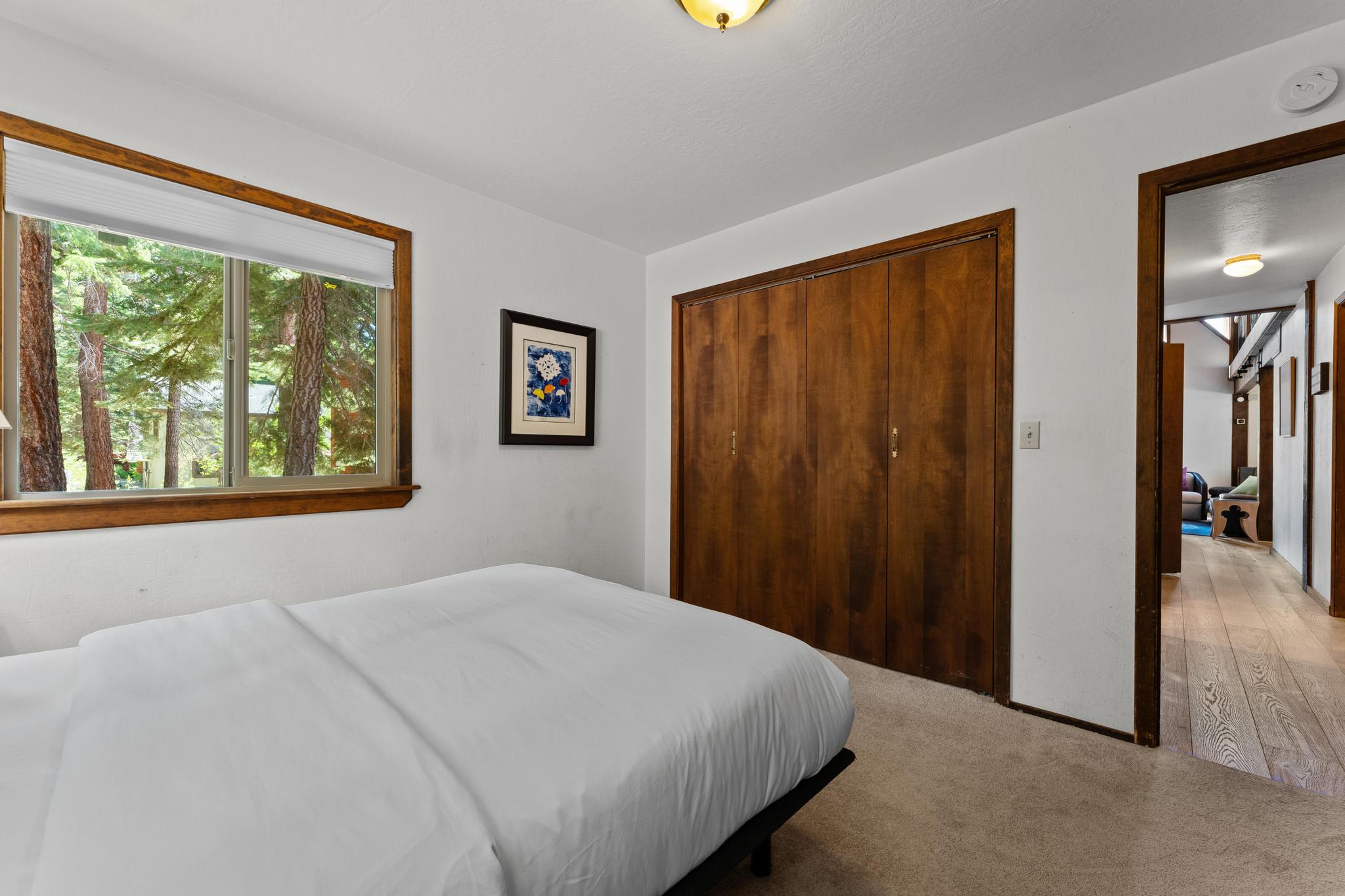 Bedroom in a Tahoe Vista vacation rental with a white bed, wooden closet, window view of trees, and hallway.