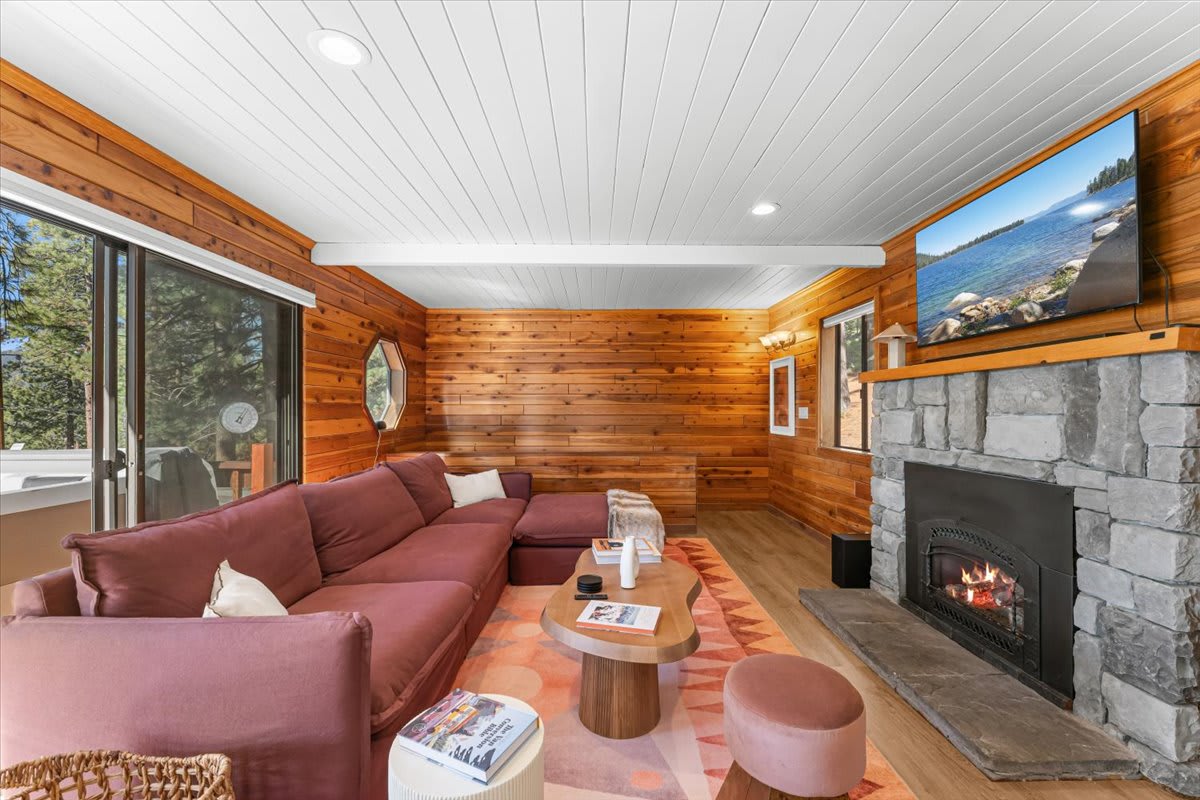 Cozy Kings Beach vacation rental living room with wooden walls, fireplace, large sofa, and scenic nature view through windows.